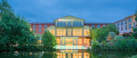 A hotel with a pool, surrounded by greenery, and a reflection in the water.