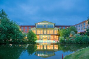 A hotel with a pool, surrounded by greenery, and a reflection in the water.