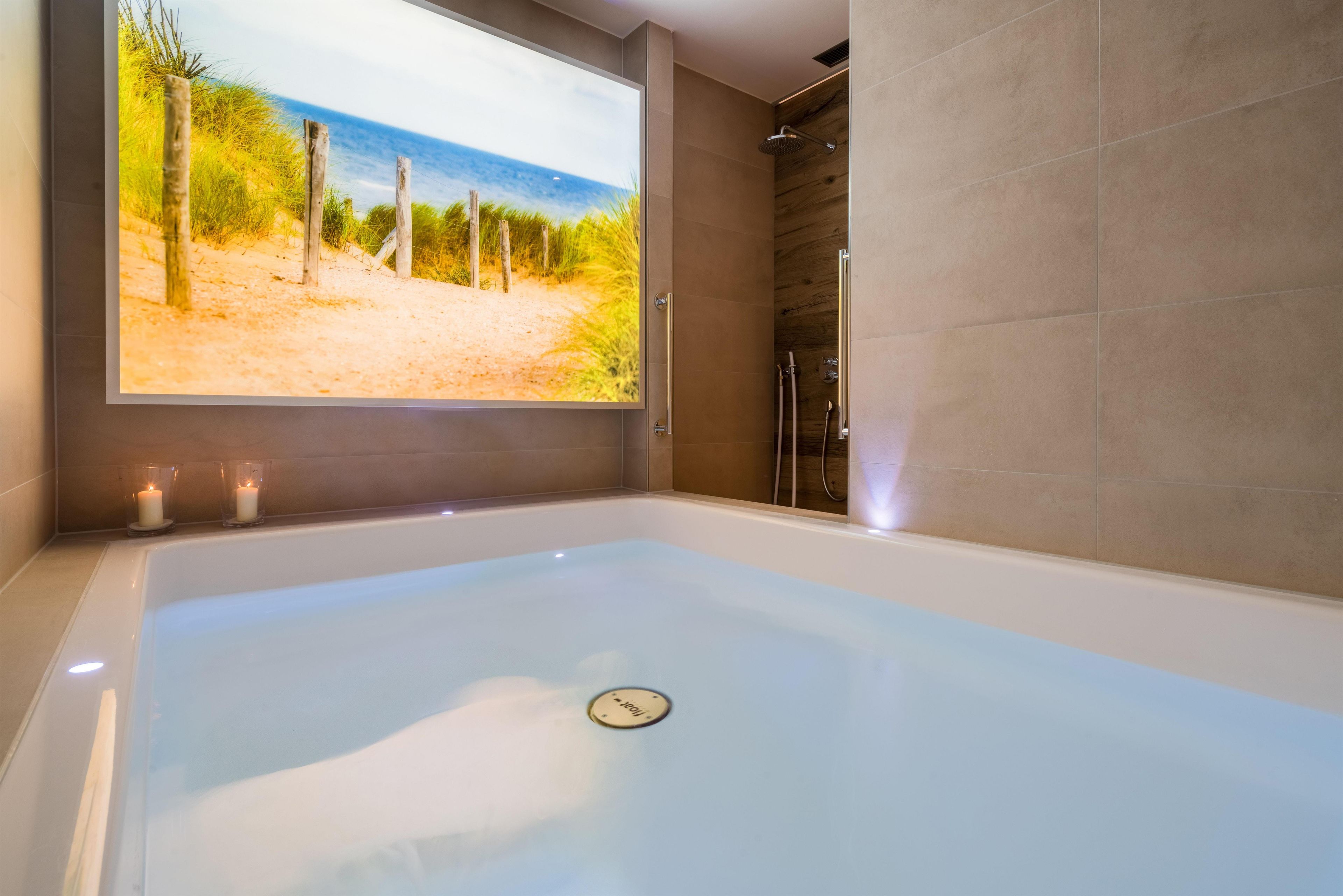A modern bathroom with a large bathtub, a shower, and a view of a beach through a window.