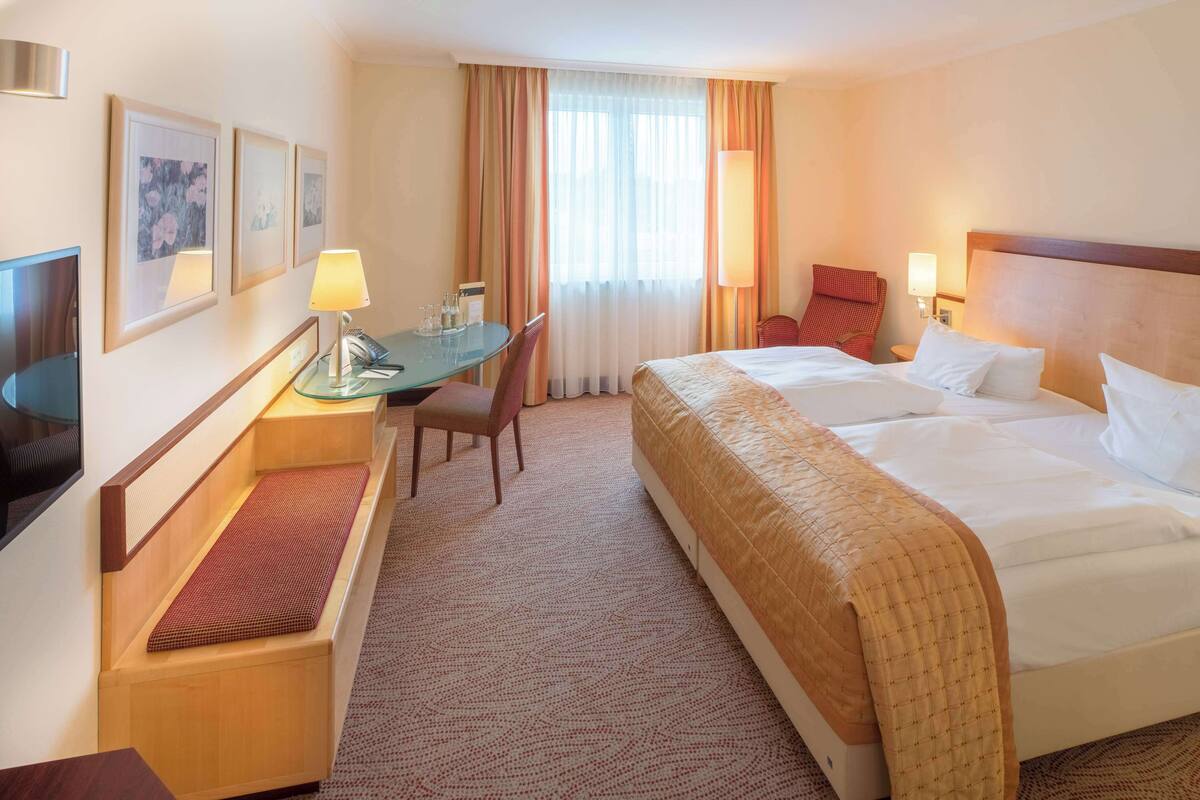Ein Hotelzimmer mit einem Bett, einem Schreibtisch mit Stuhl, einem Fernseher und einem Fenster mit Vorhängen.