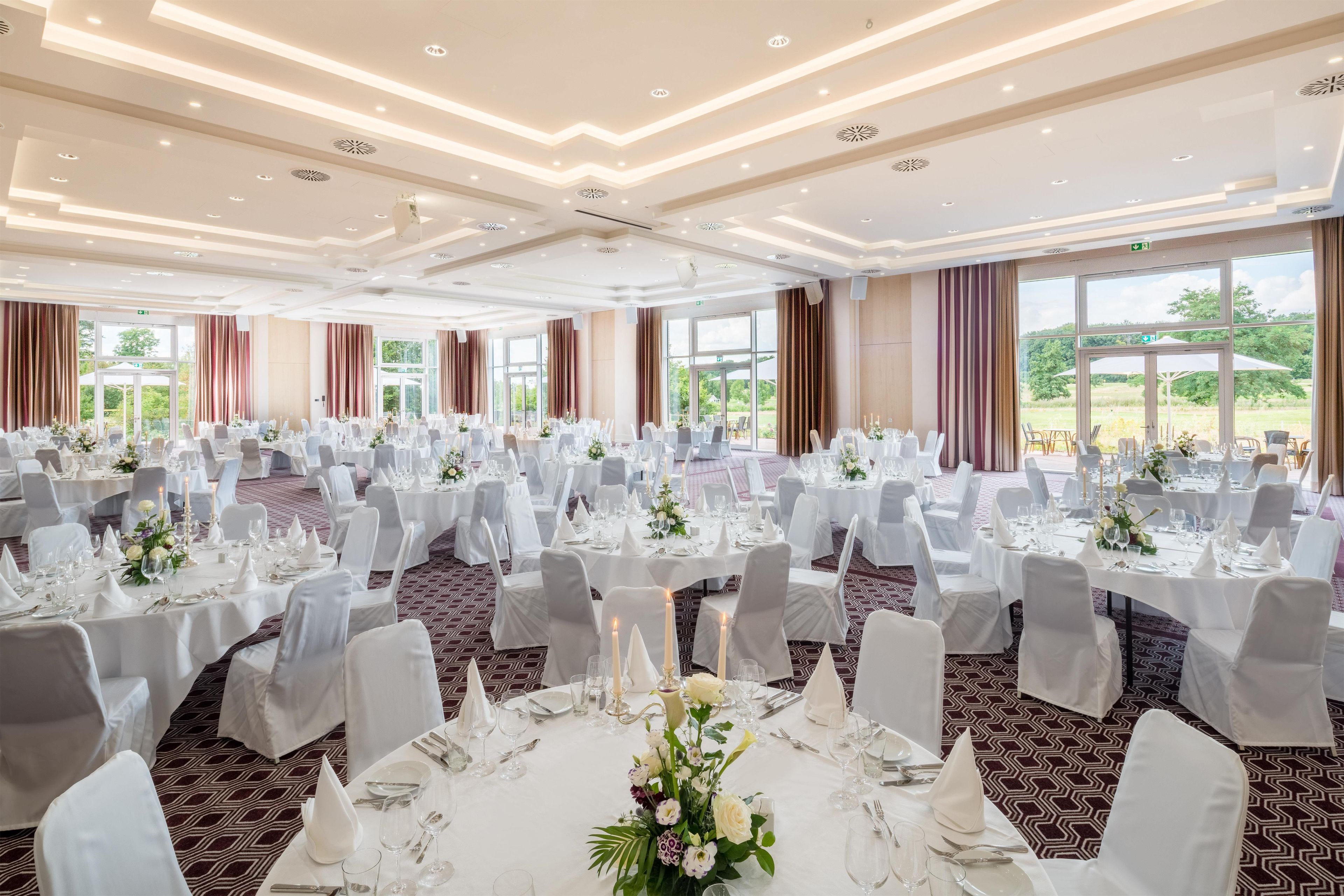 A banquet hall with round tables set for dining, large windows offering a view of the outdoors, and a modern ceiling design.
