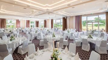 A banquet hall with round tables set for dining, large windows offering a view of the outdoors, and a modern ceiling design.