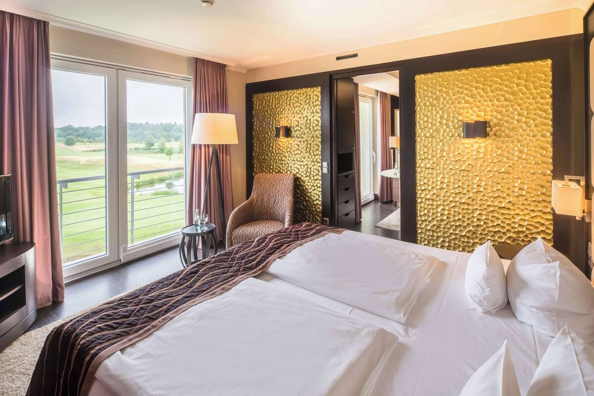 Ein Hotelzimmer mit einem großen Bett, einem Sessel, einem Fernseher und Blick auf einen Golfplatz durch die Fenster.