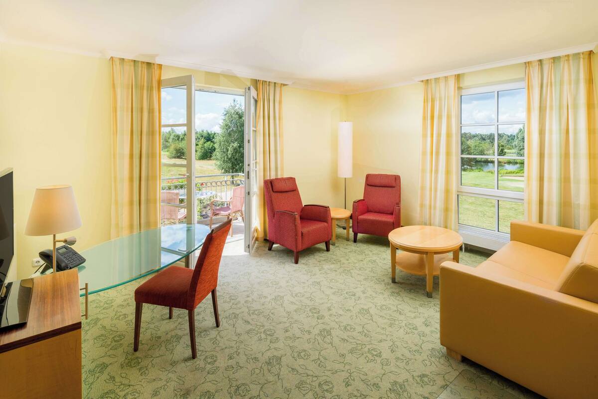 Ein Hotelzimmer mit Glastisch, zwei roten Sesseln, einem gelben Sofa und Blick auf einen Garten durch große Fenster.