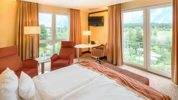 Ein Hotelzimmer mit einem Bett, zwei Sesseln, einem kleinen Tisch, einem Fernseher und großen Fenstern mit Blick auf Grünflächen.