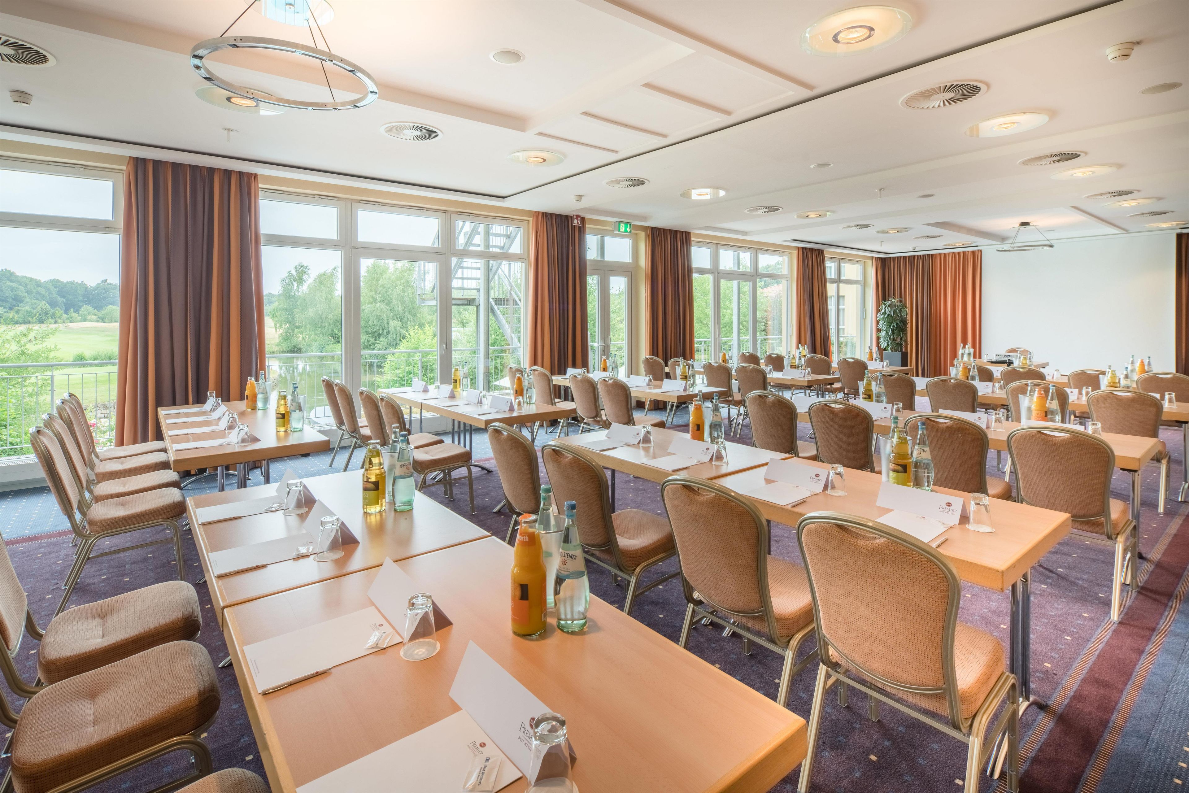 Un salón de conferencias con varias mesas y sillas, amplias ventanas con cortinas y vistas al exterior con vegetación.