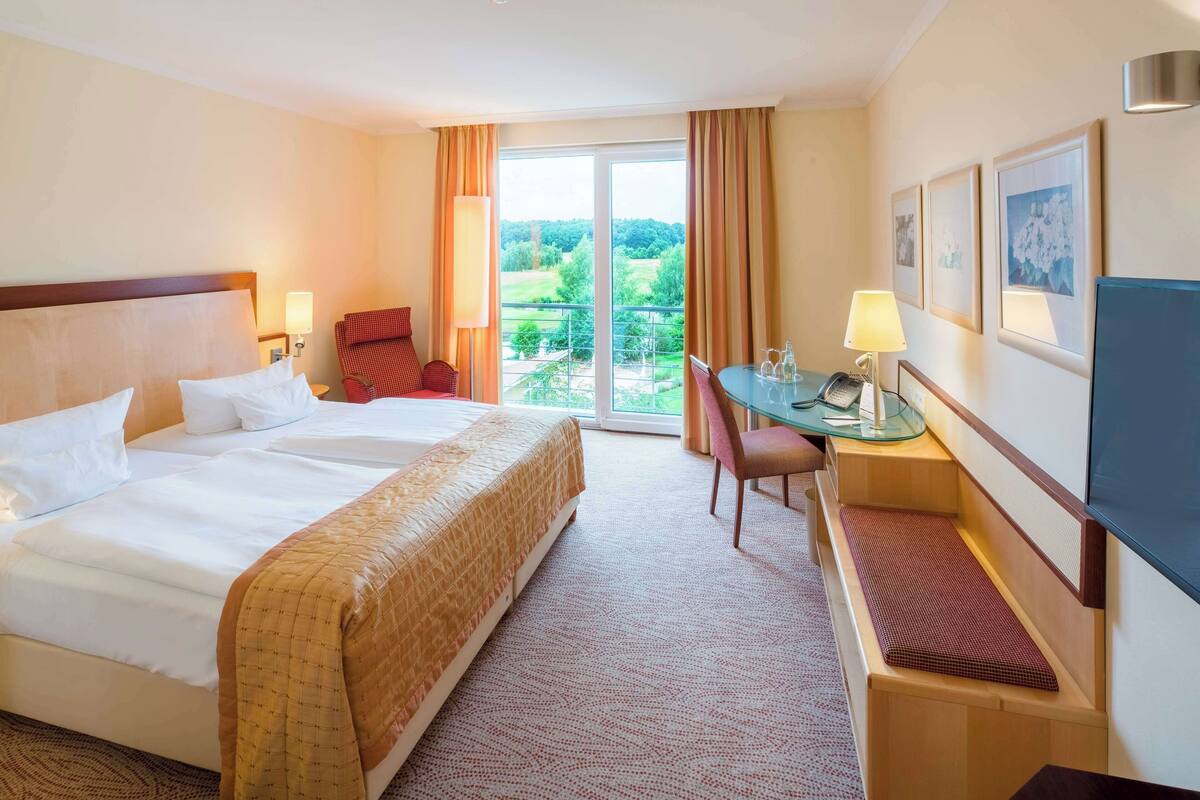 Ein Hotelzimmer mit einem großen Bett, einem Schreibtisch mit Stuhl, einem Fernseher und einem Balkon mit Blick auf Grünflächen.