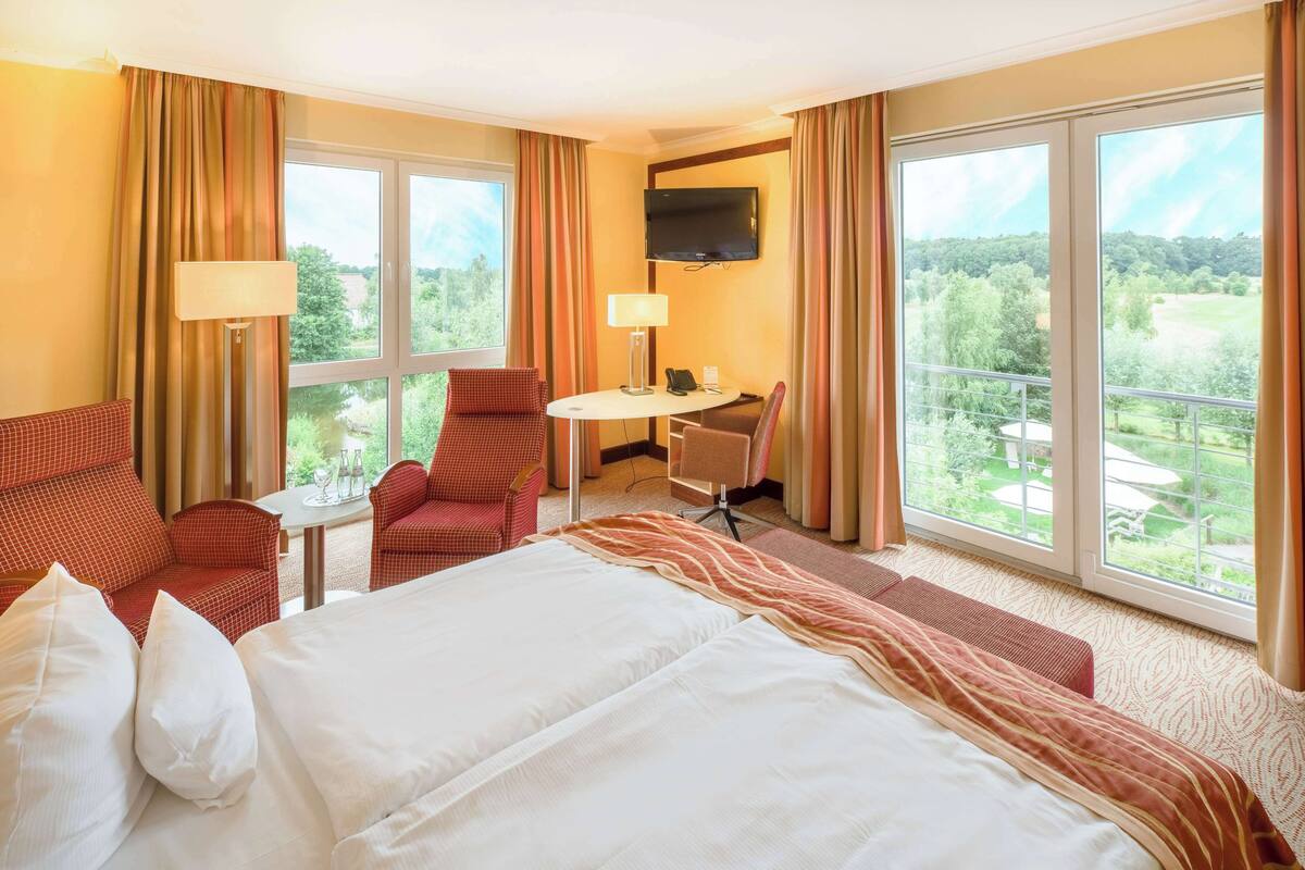 Ein Hotelzimmer mit einem großen Bett, zwei Sesseln, einem kleinen Tisch, einem Fernseher und großen Fenstern mit Blick auf Grünflächen.