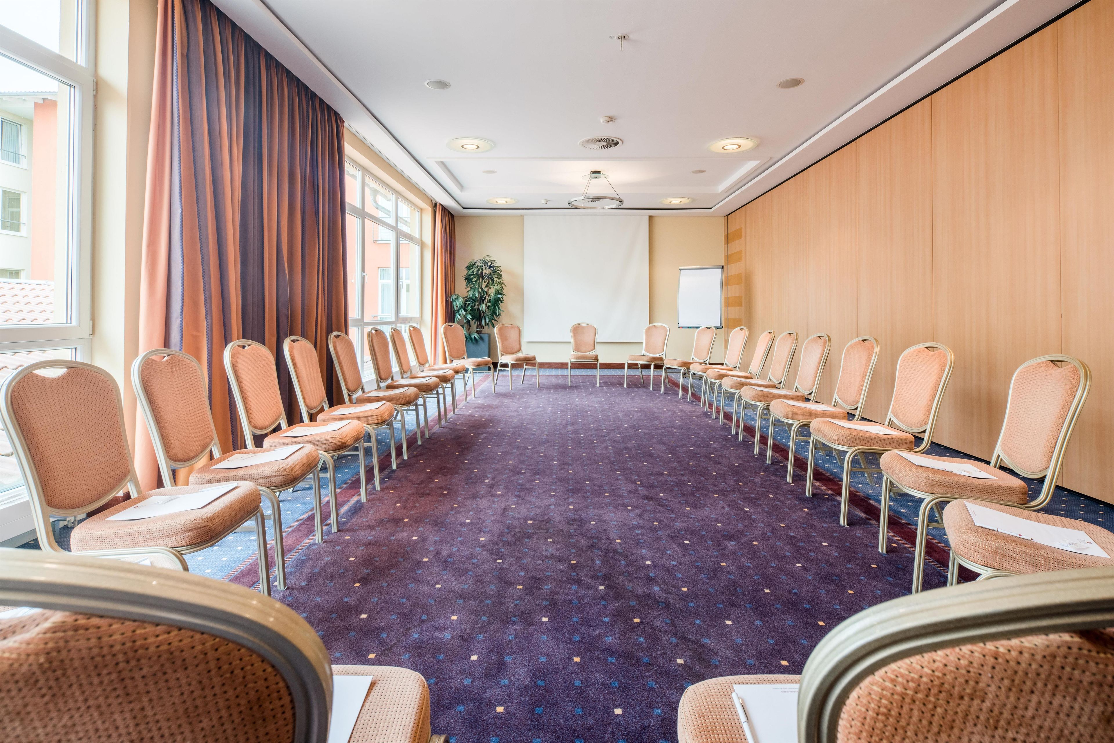 Un salón de conferencias con una larga mesa, sillas, ventanas amplias y una planta.