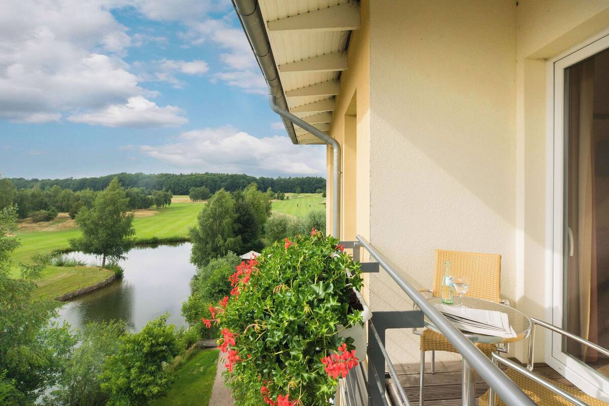 Ein Balkon mit Blick auf einen See und einen Golfplatz, ein Tisch mit einem Stuhl sowie eine hängende Pflanzampel mit Blumen.