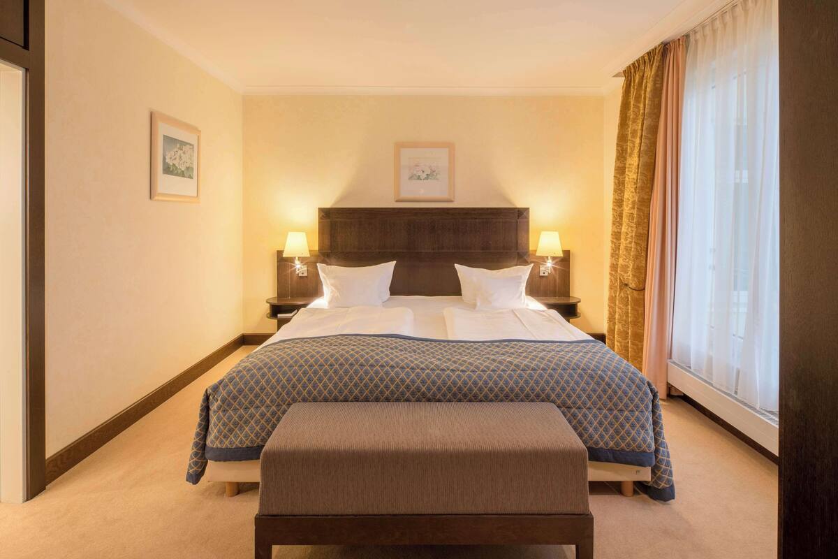 Ein Hotelzimmer mit einem großen Bett, zwei Nachttischlampen, einem Kopfteil, einem Fenster mit Vorhängen und einem gerahmten Bild an der Wand.
