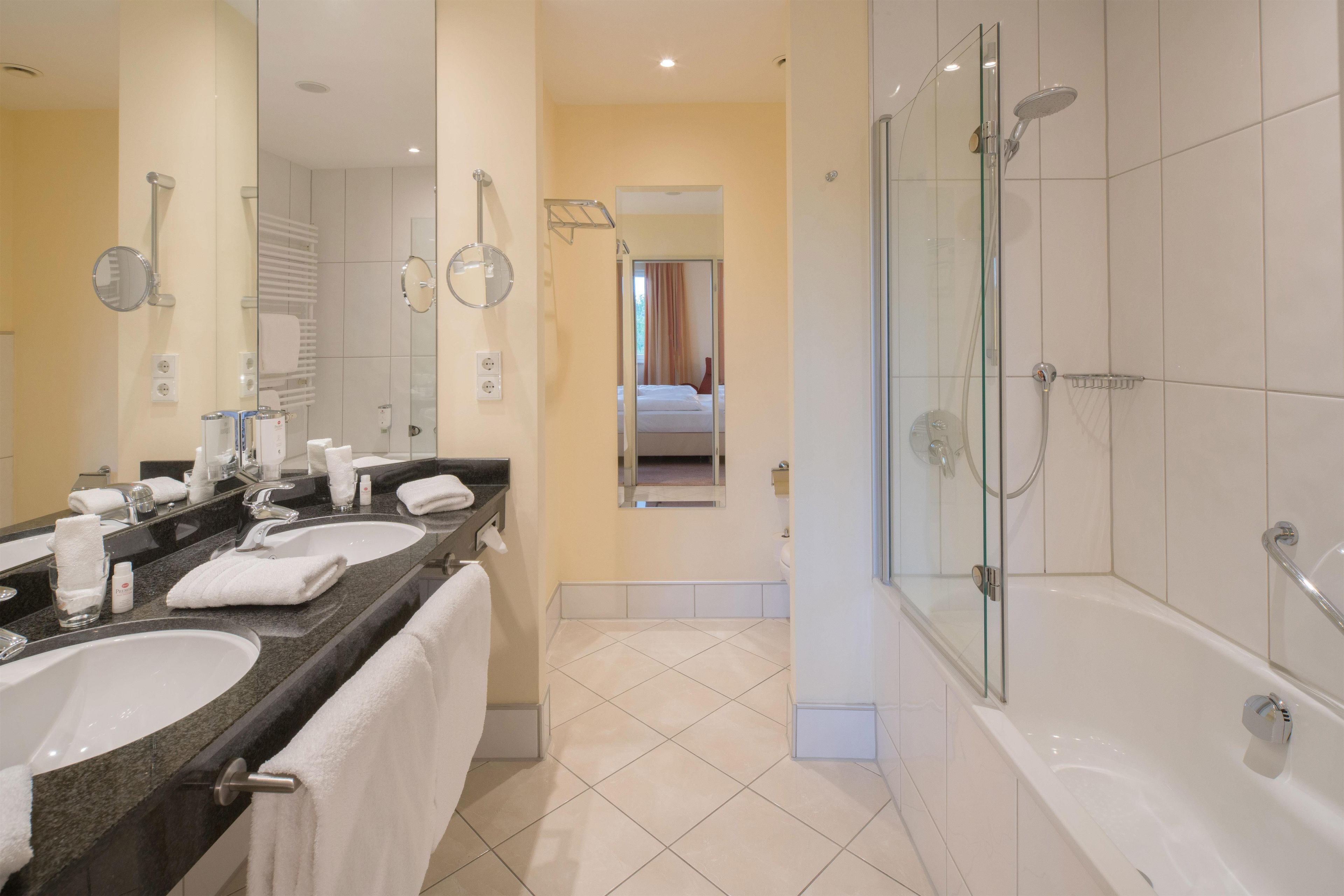 Un baño moderno con lavamanos dobles, un espejo grande, cabina de ducha de vidrio y una habitación visible al fondo.