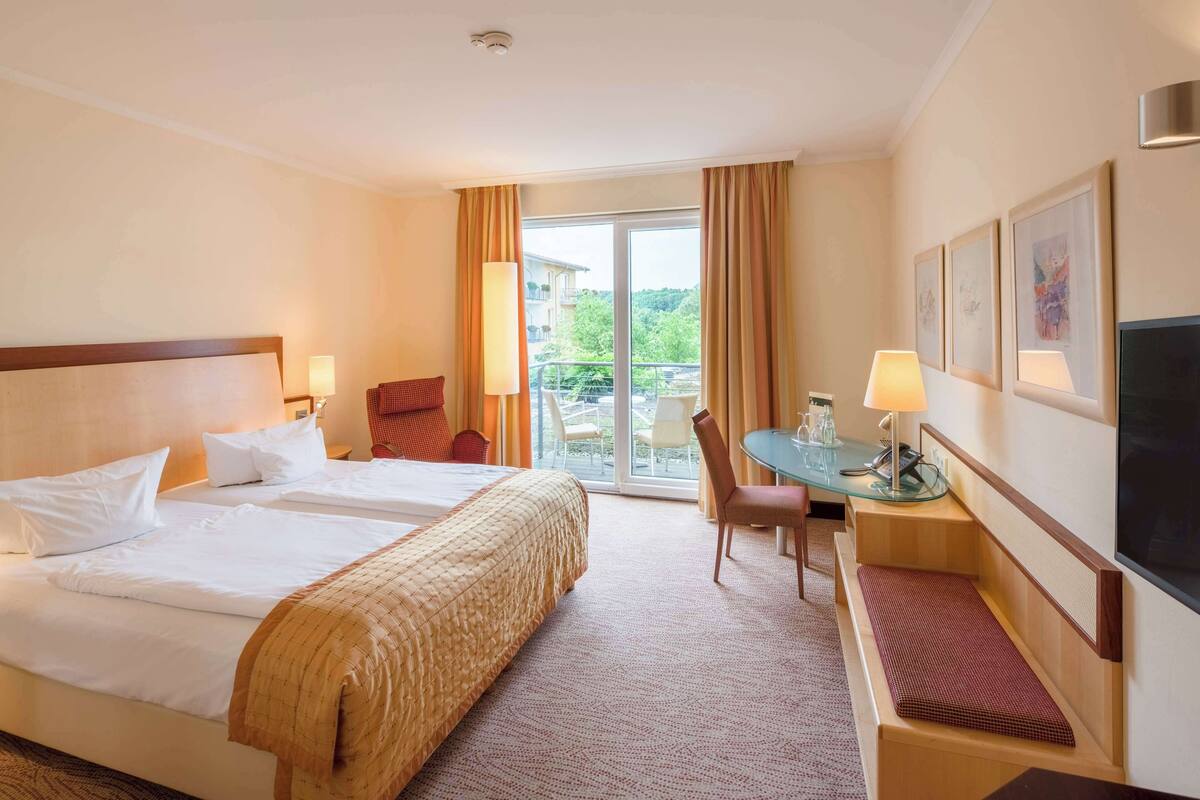 Ein Hotelzimmer mit zwei Betten, einem Schreibtisch mit Stuhl, einem Fernseher und einem Balkon mit Vorhängen.