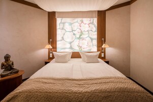 Un dormitorio con una cama, dos lámparas de noche, una estatua y una ventana con motivos florales.