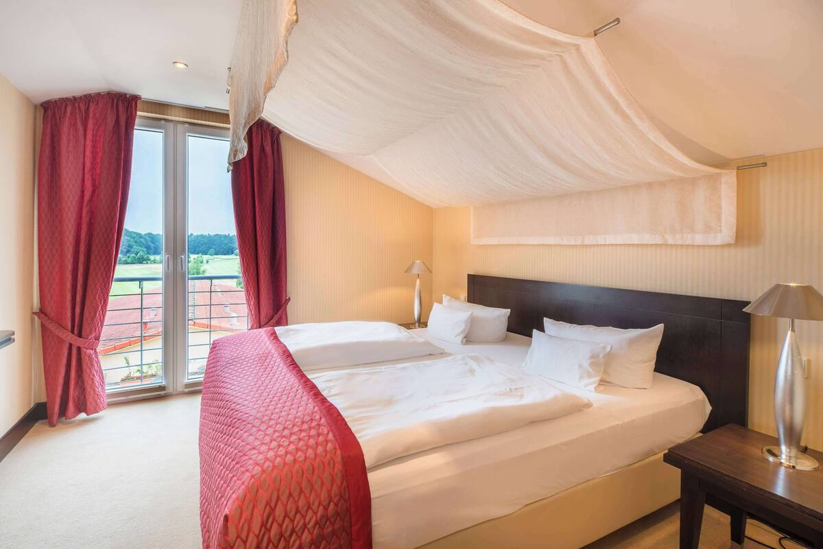 Ein Hotelzimmer mit einem großen Bett, einem Nachttisch, einer Lampe und Blick auf eine grüne Landschaft durch das Fenster.