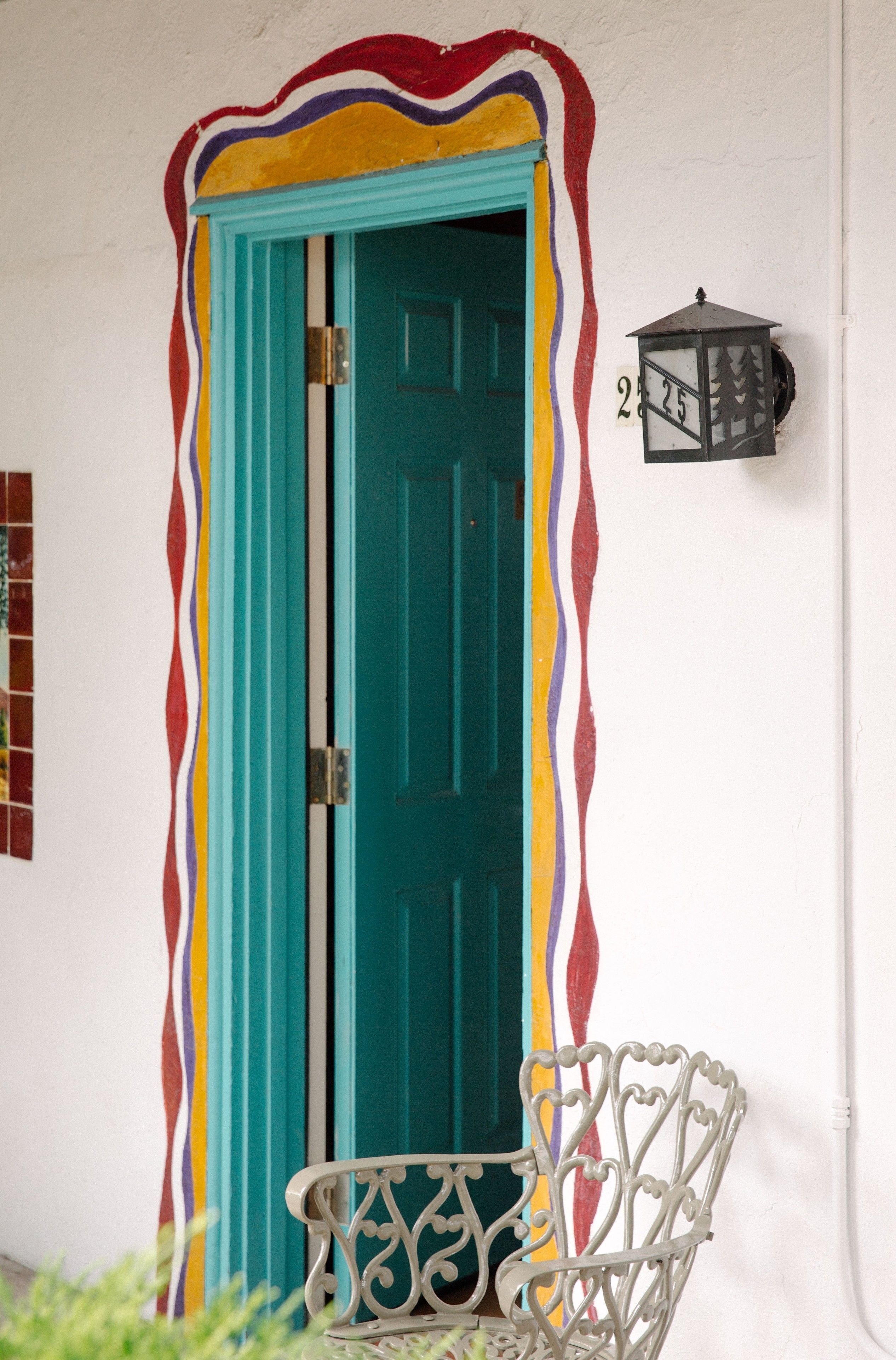 Una puerta turquesa con un diseño ondulado y colorido, una silla blanca con detalles de metalurgia, y un farol adosado a la pared con el número 225.