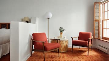 Una habitación con dos sillas rojas, una mesita redonda con un florero y una cama con ropa de cama blanca.