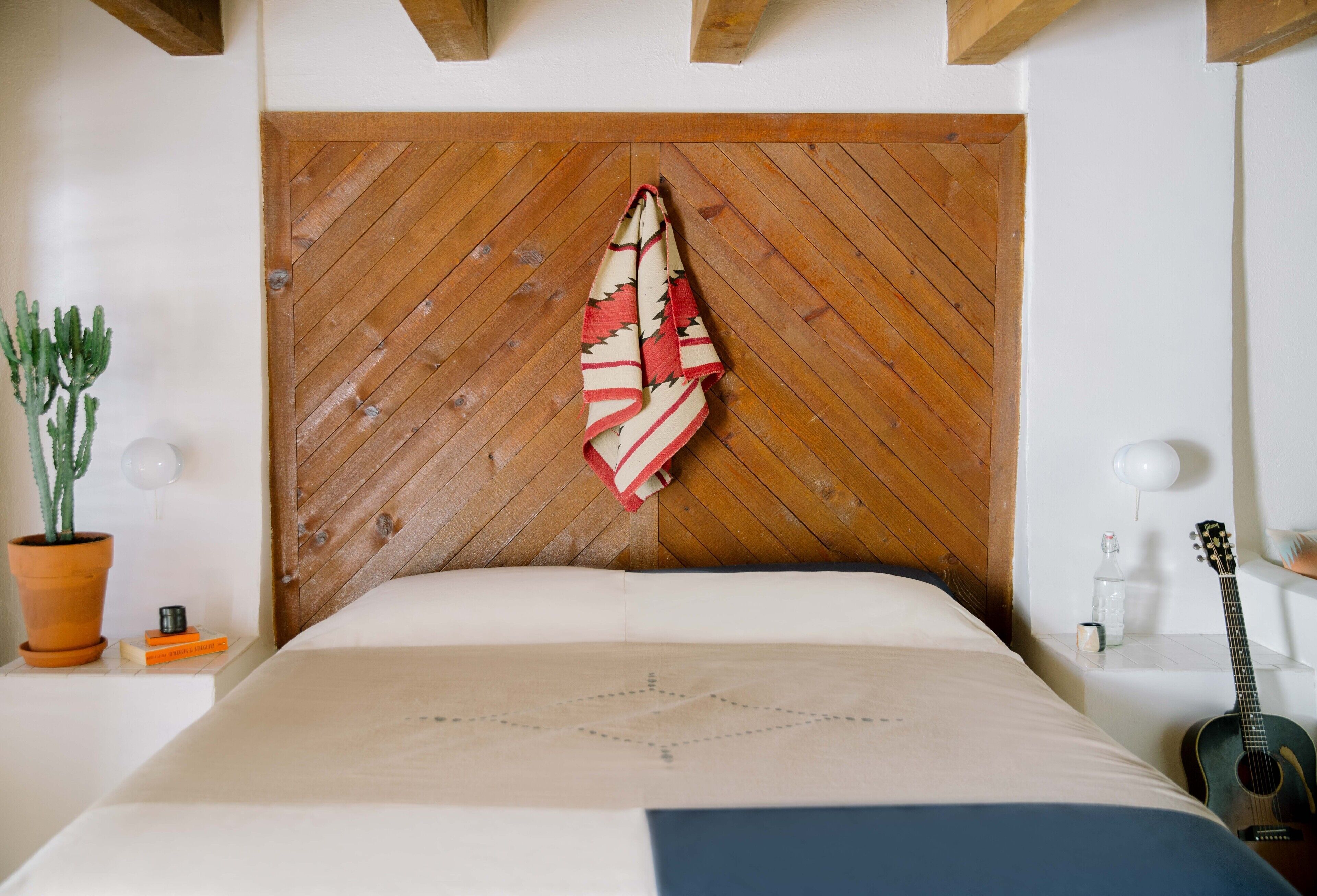 A bedroom with a wooden headboard, a bed with a geometric-patterned bedspread, a potted plant, a guitar, and a small table with a bottle and a candle.