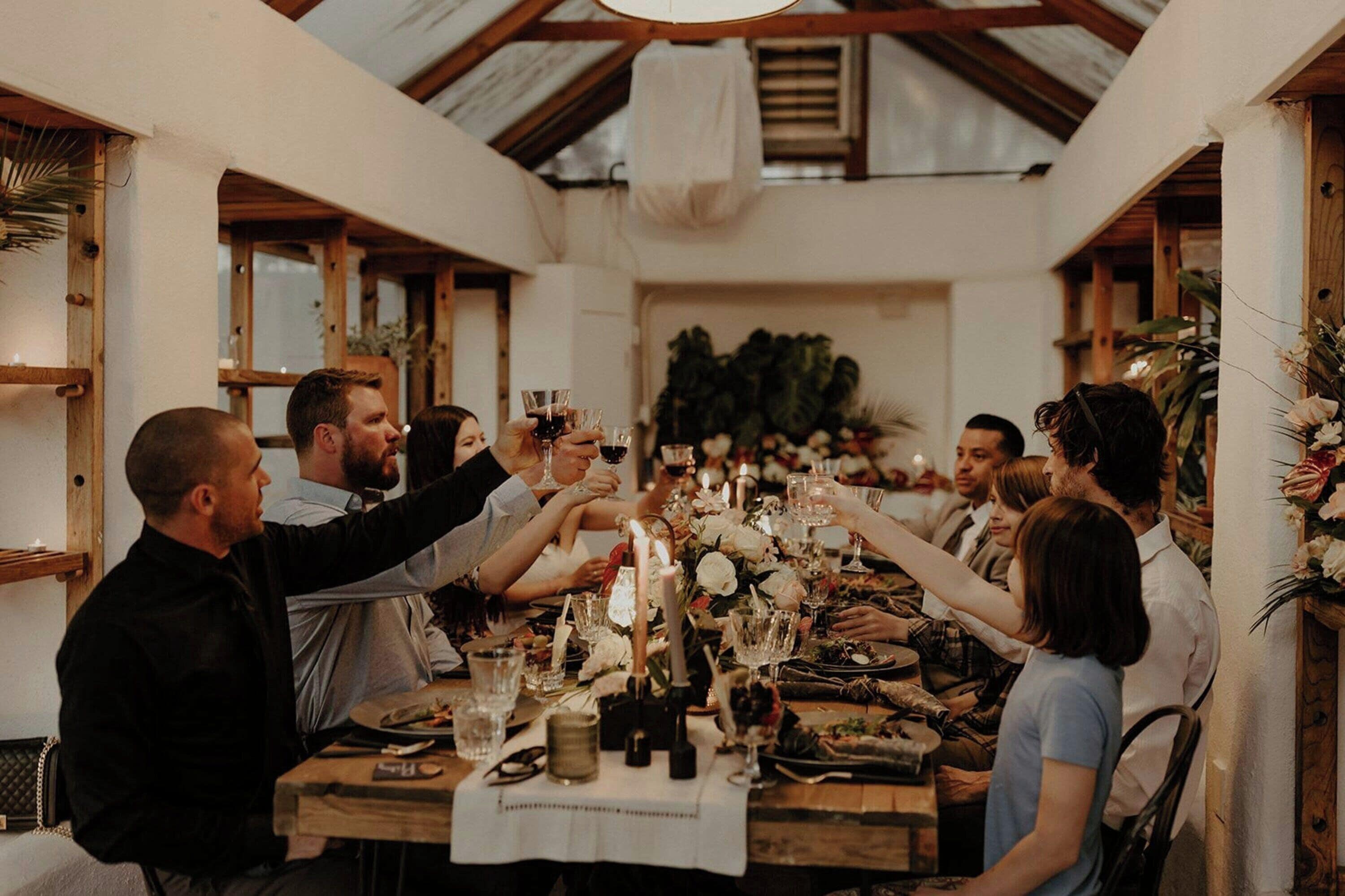 A group of people toasting at a wooden table with a white tablecloth, surrounded by floral arrangements and candles.