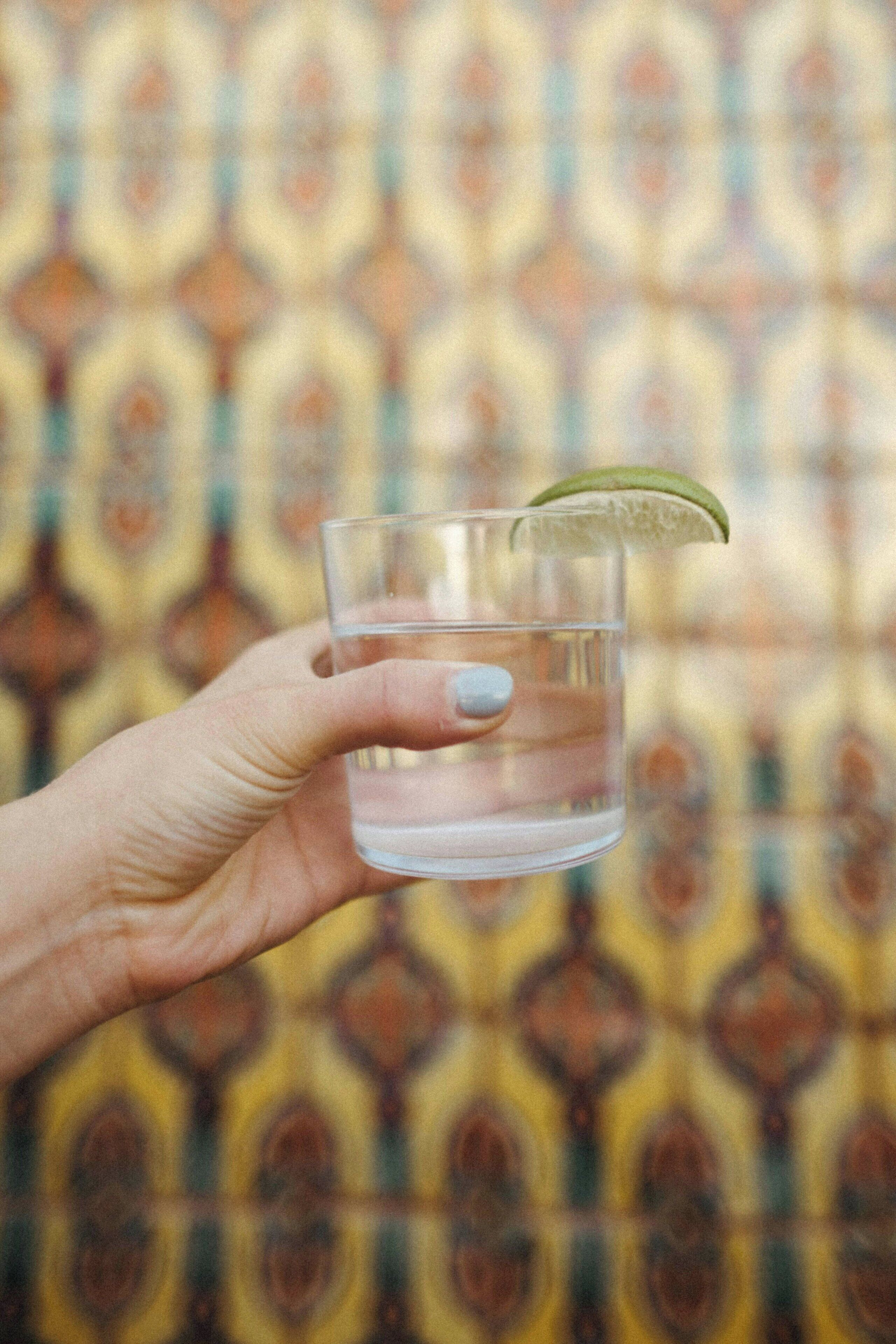 Une main tenant un verre transparent dans lequel on a mis un quartier de citron vert, le tout sur un fond à motifs.