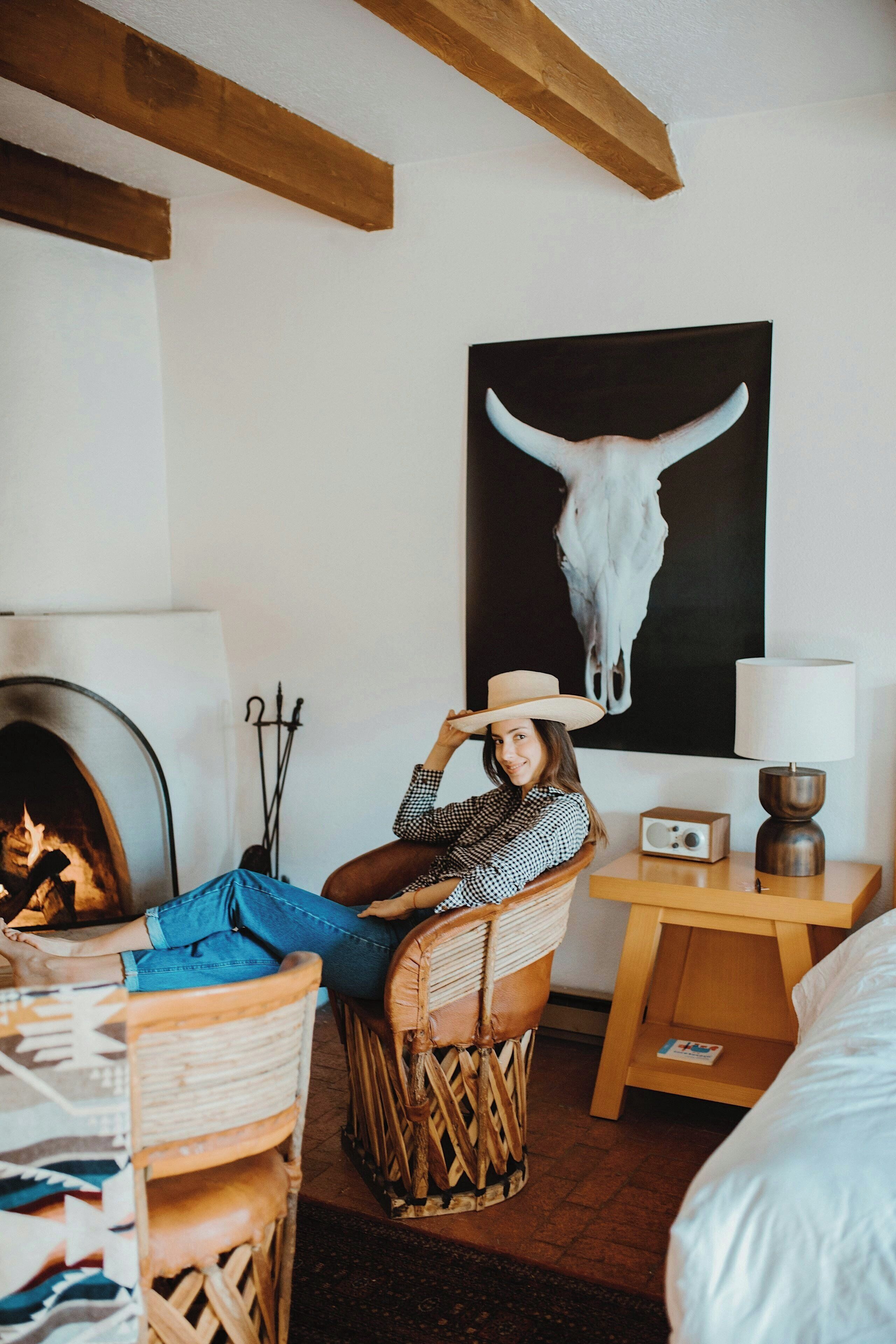 Une pièce chaleureuse avec une cheminée, une œuvre d’art représentant un crâne de vache, un fauteuil en osier et une table de chevet avec une lampe.