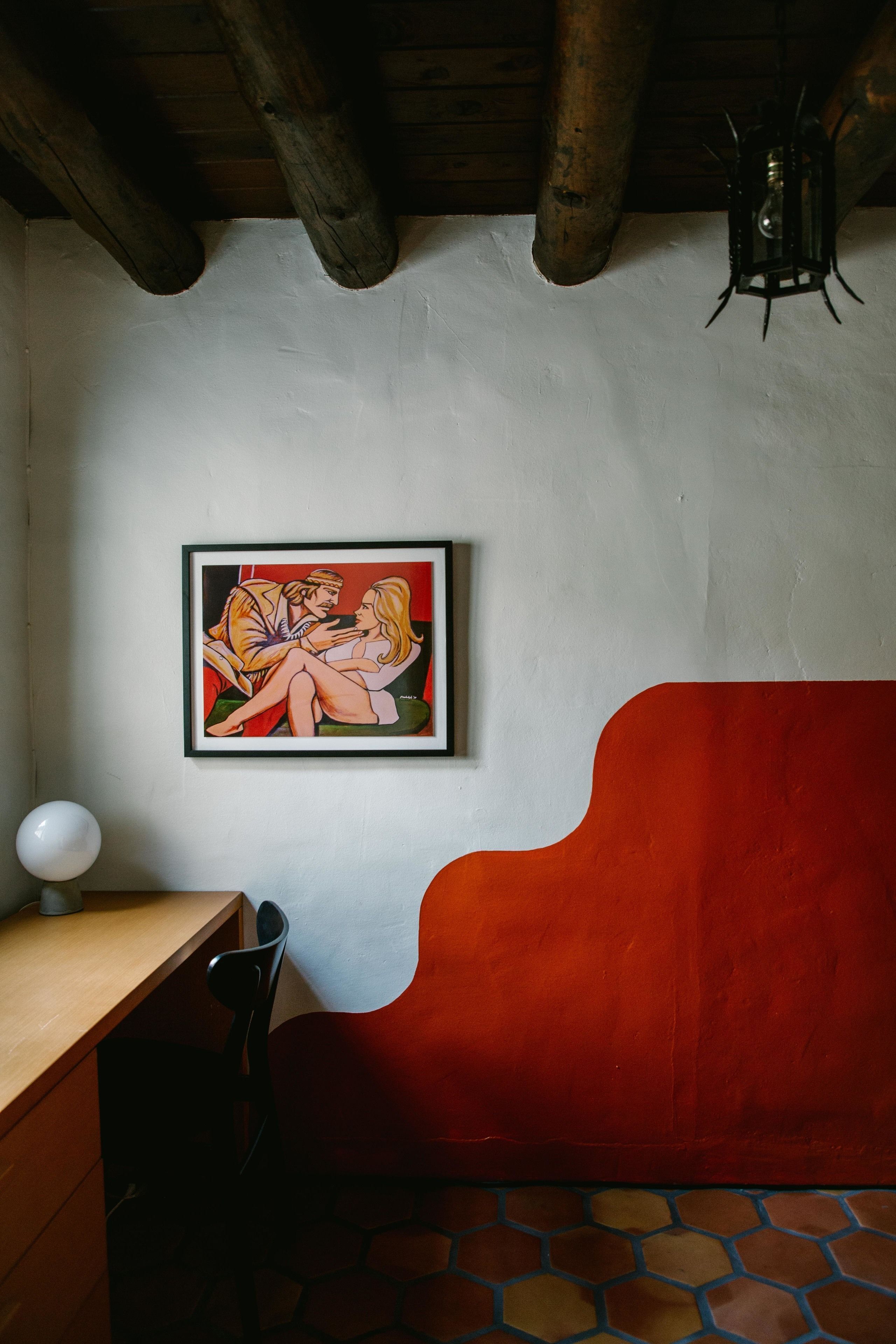 A room with a wooden ceiling, a red cushioned bench, a framed artwork on the wall, and a desk with a lamp.