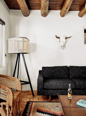 A living room with a black sofa, a woven floor lamp, a rustic wooden ceiling, a cow skull wall decoration, and a coffee table with a bottle and book. - El Rey Court (Santa Fe)