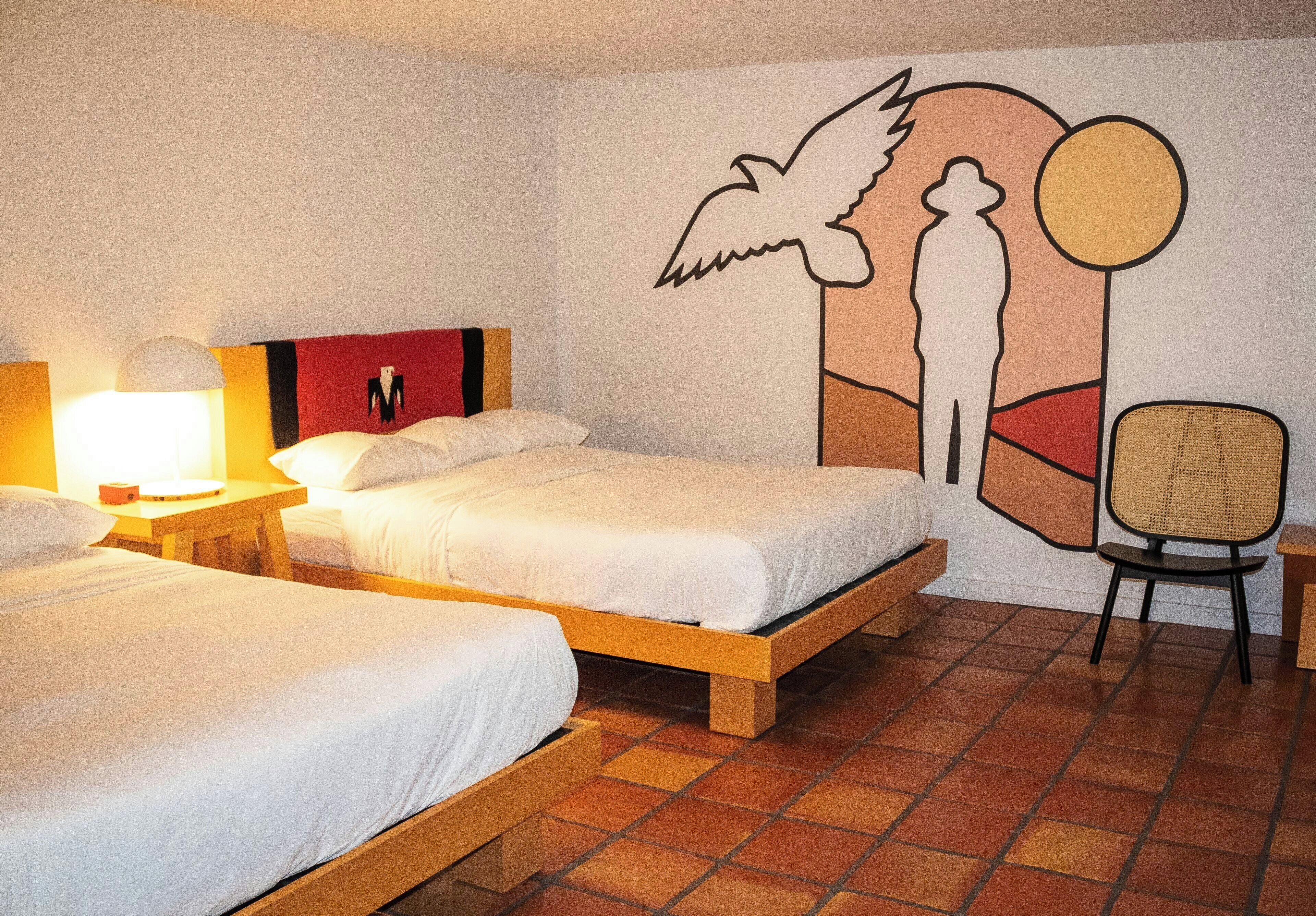 Habitación de hotel con dos camas, un mural en la pared, una silla y una lámpara.