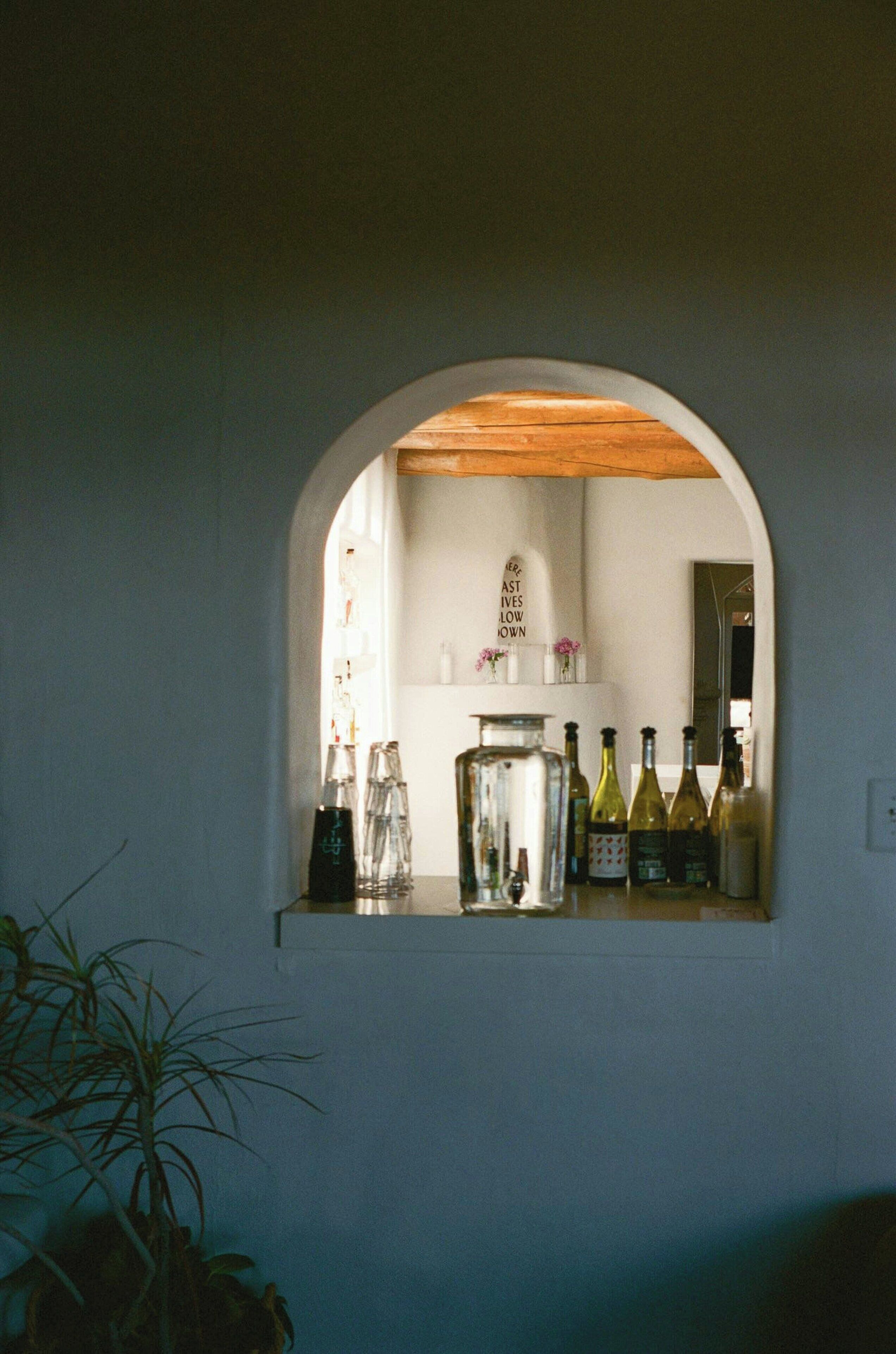 Una zona de bar con techo de madera y pared blanca. Hay botellas y vasos sobre la barra.