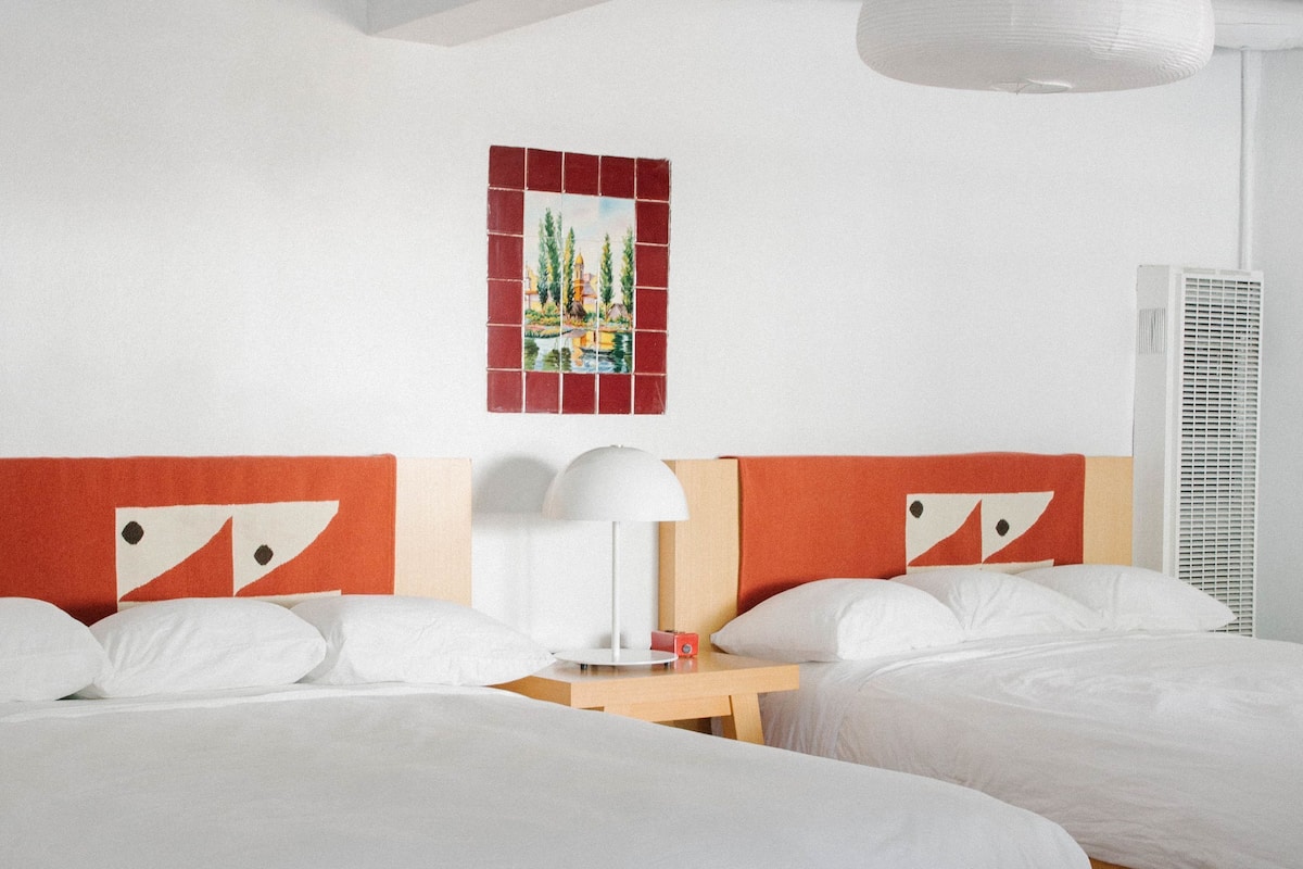 A hotel room with two beds, a nightstand, a lamp, and a wall art piece.