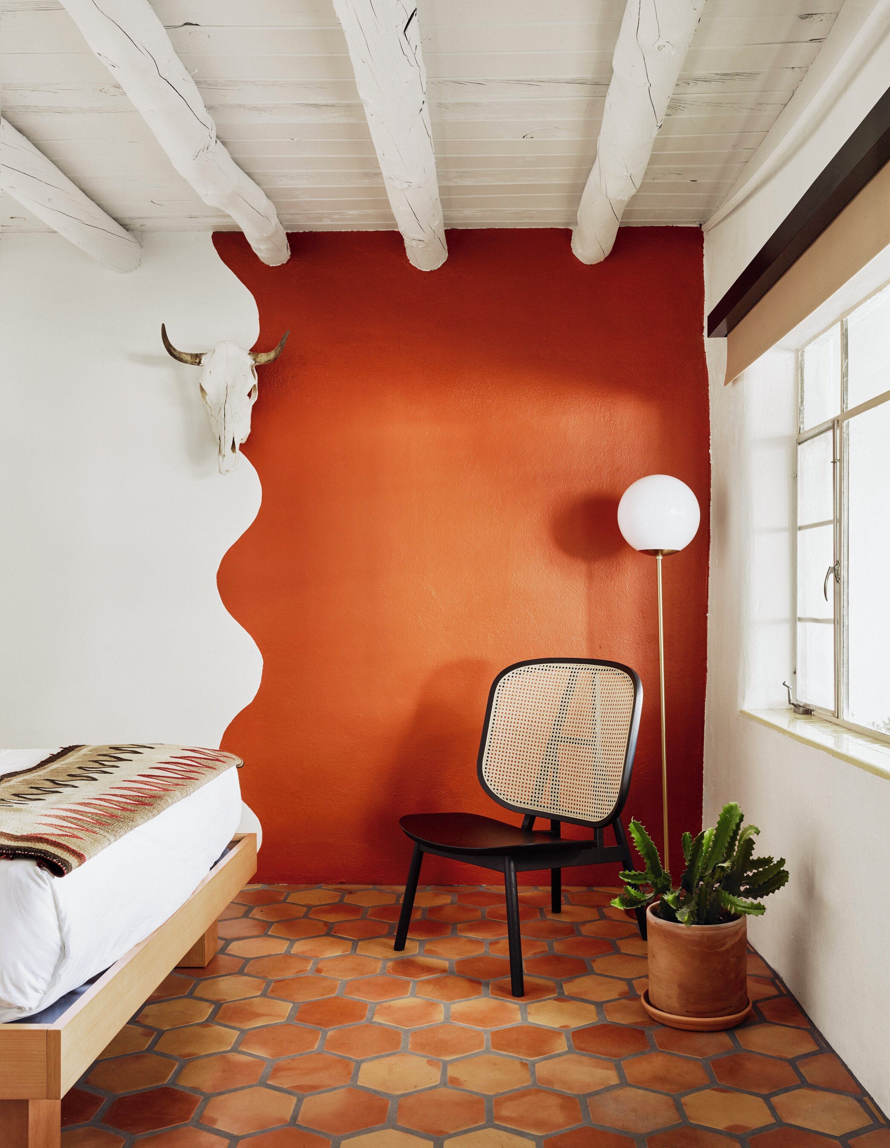 A small room with a red wall, a white ceiling, a wooden bed, a black chair, a floor lamp, and a potted plant.