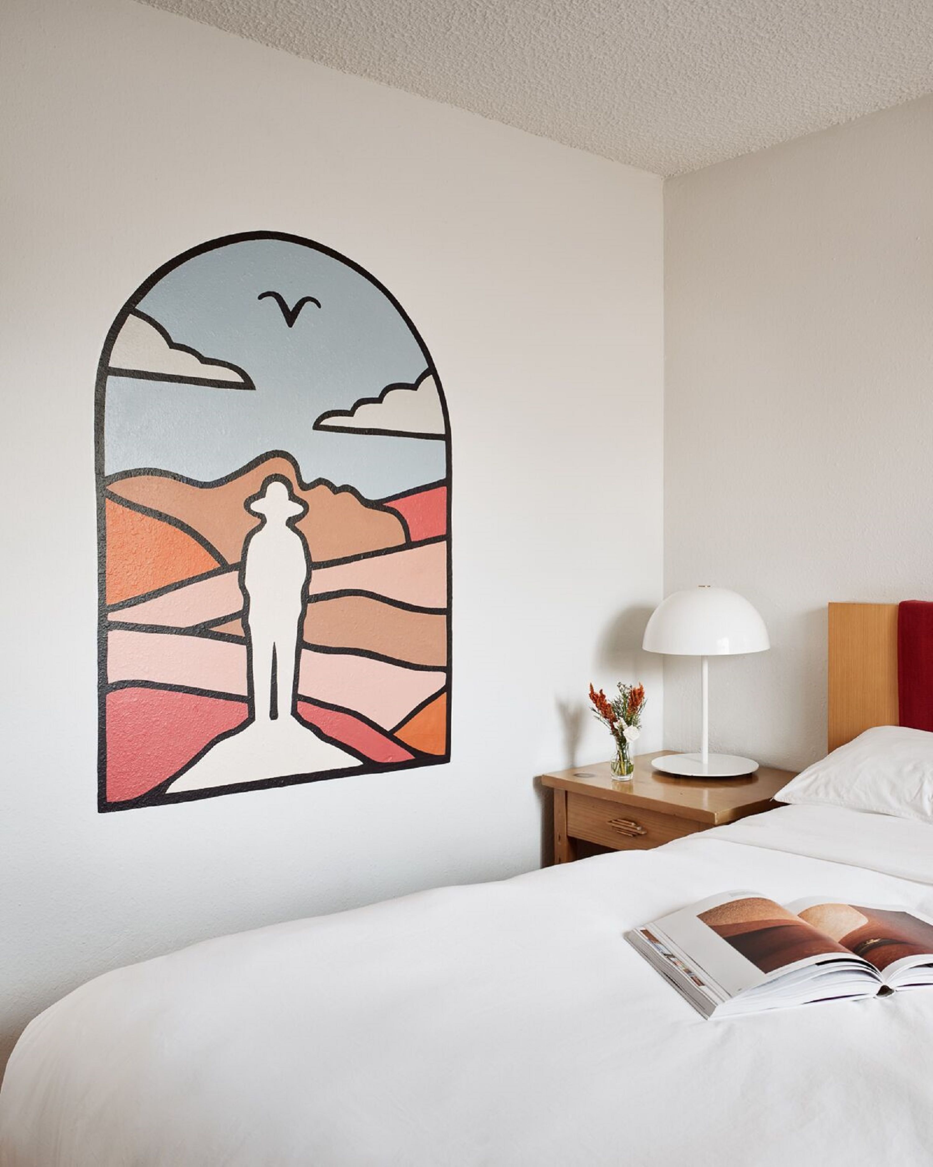 Una habitación de hotel con cama, mesita de noche con lámpara, y una pared decorada con arte abstracto y colorido.