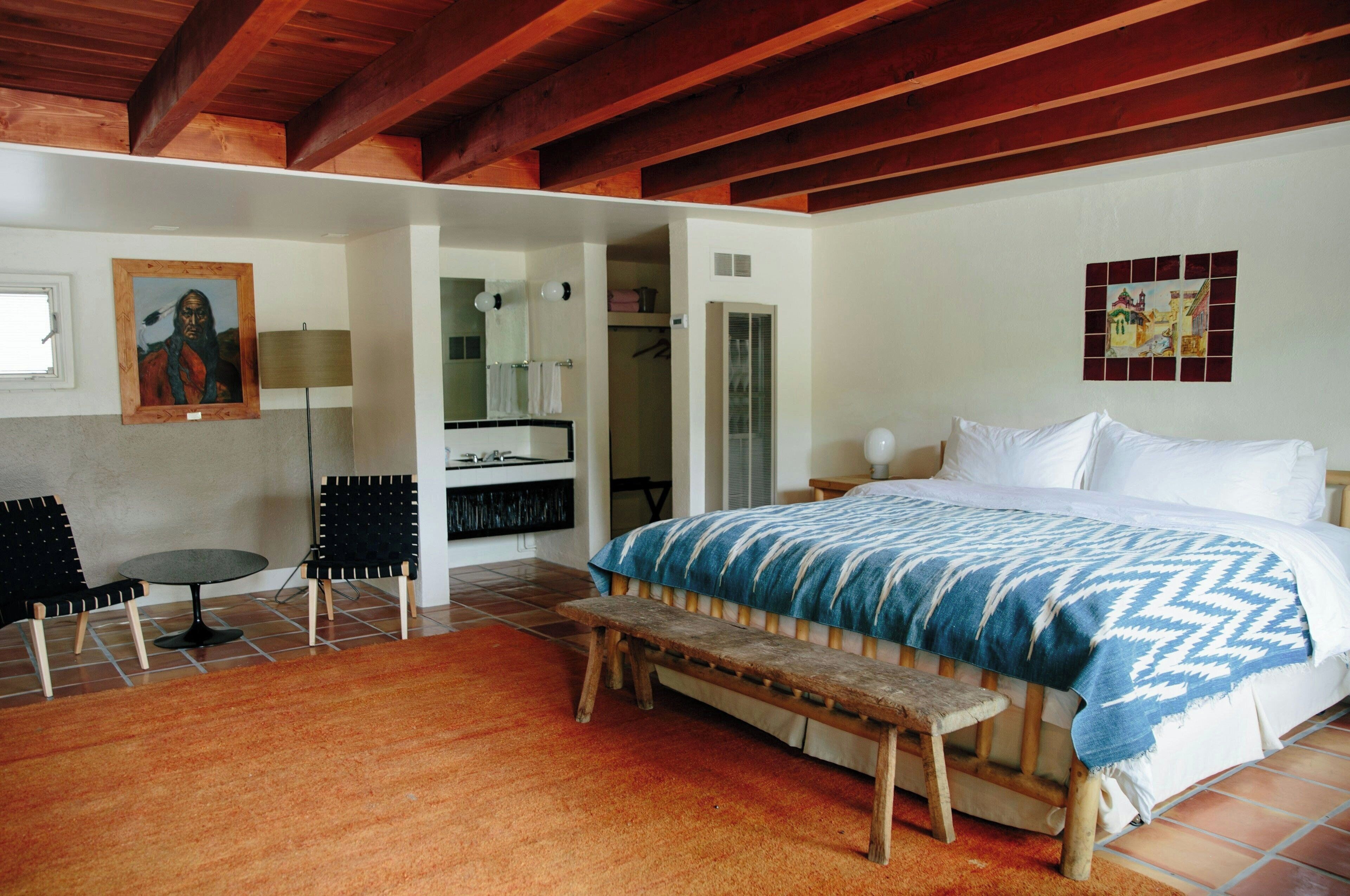 A bedroom with a bed, a bench, a chair, a small table, and a painting on the wall.