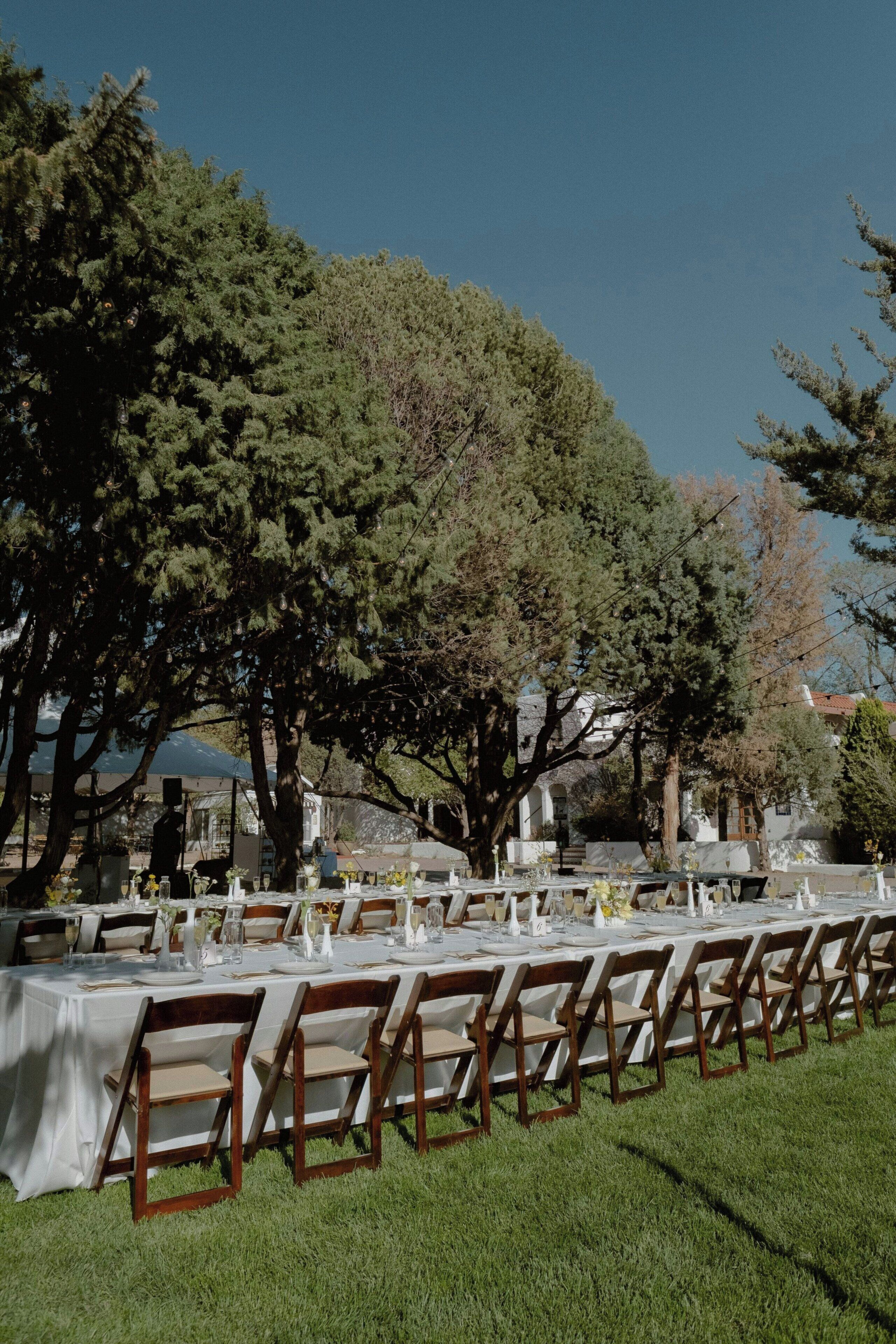 An outdoor dining area with tables set for guests, surrounded by trees and a clear sky.