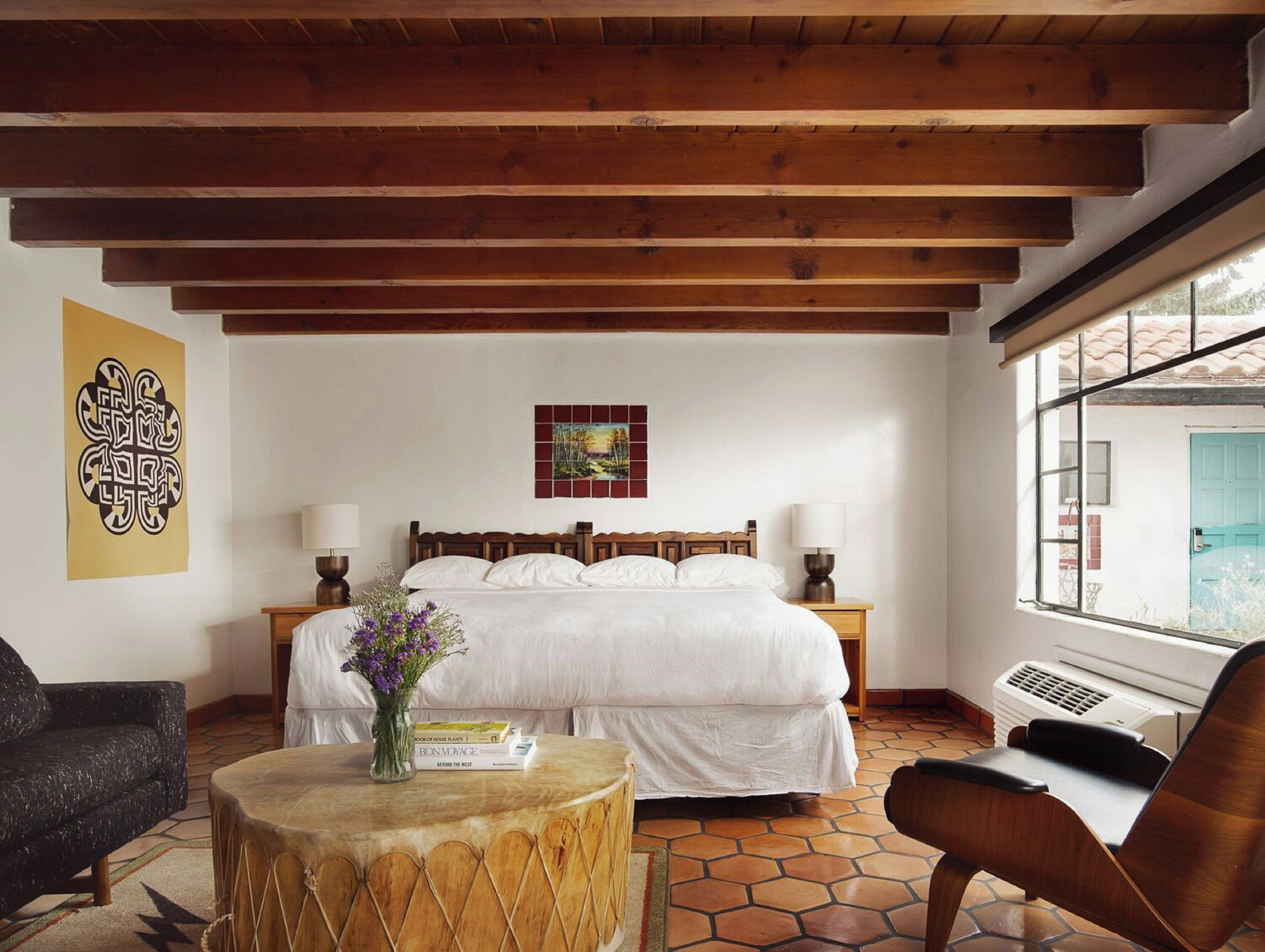 A bedroom with a wooden ceiling, a bed with white bedding, a window with a view, and a round wooden table with a vase.