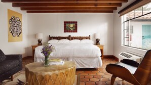 A bedroom with a large bed, wooden beams, a window with a view, a bedside table with a lamp, and a chair. - El Rey Court (Santa Fe)
