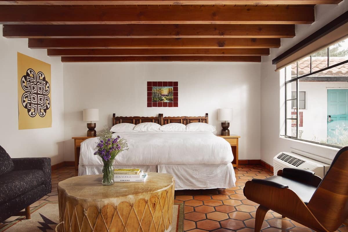 A bedroom with a large bed, wooden beams, a window with a view, a bedside table with a lamp, and a chair.