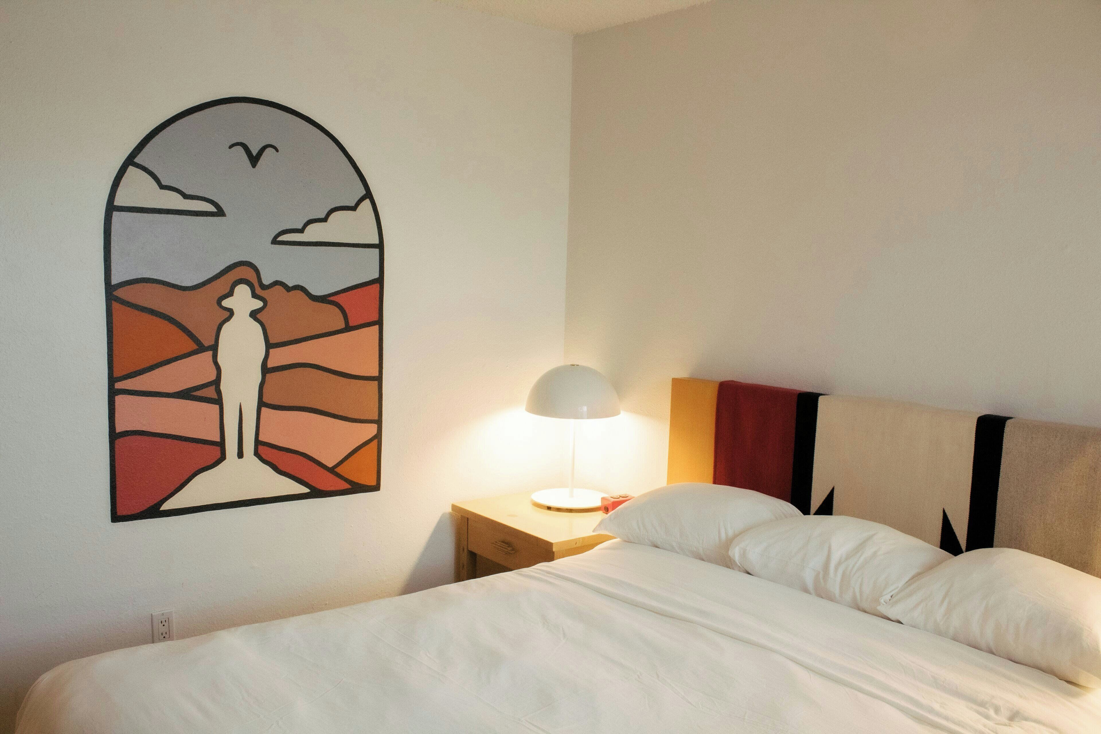 Une chambre d’hôtel comprenant un lit, une table de chevet avec une lampe, une œuvre d’art murale et une tête de lit ornée d’un motif géométrique.