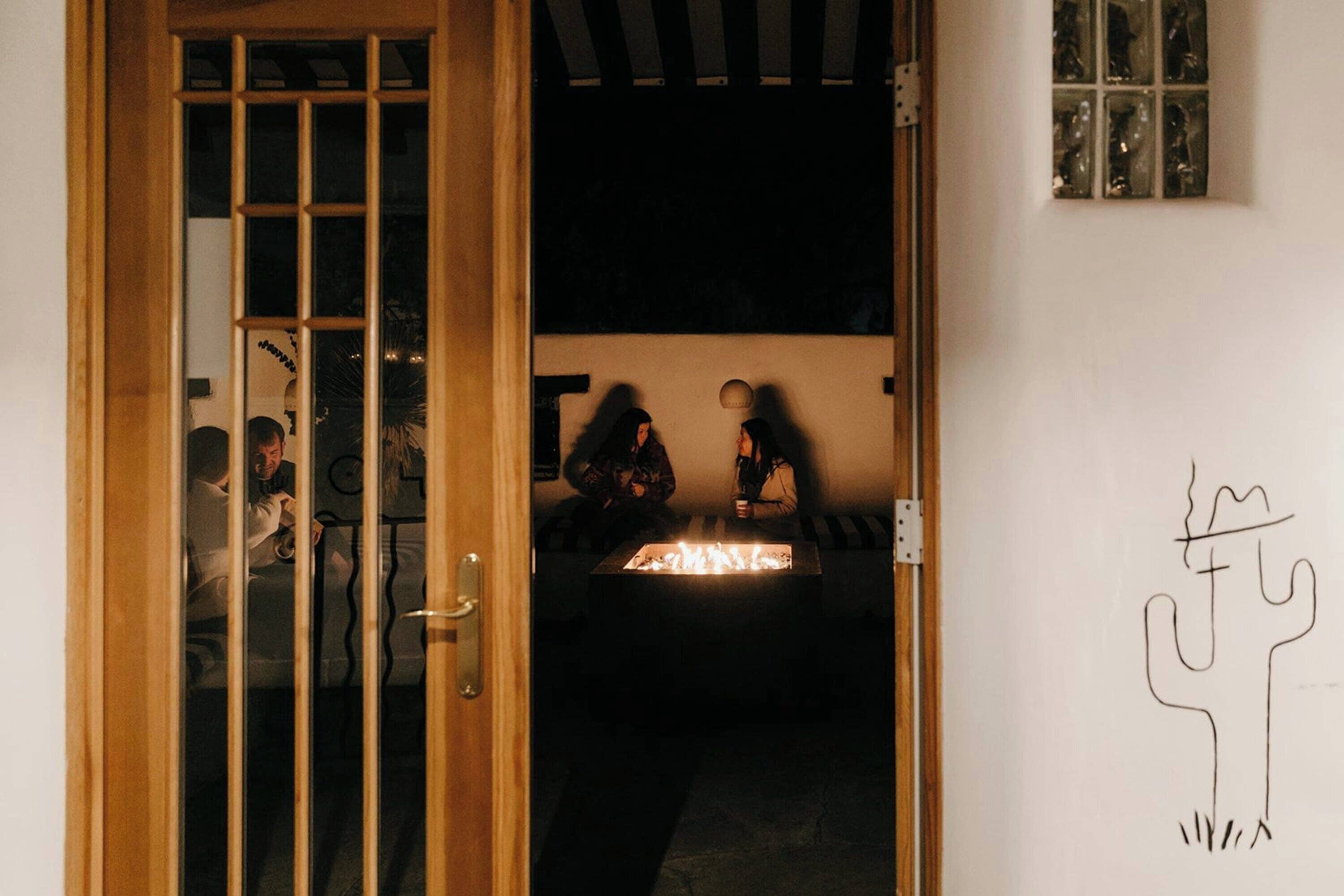 A room with a wooden door, a small table with a light source, and a wall with a simple line drawing.