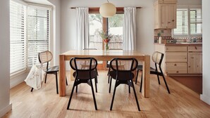 A dining area with a wooden table, four chairs, a hanging light fixture, and a view of the outdoors through a window with white curtains. - El Rey Court (Santa Fe)
