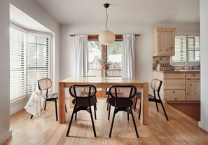 A dining area with a wooden table, four chairs, a hanging light fixture, and a view of the outdoors through a window with white curtains. - El Rey Court (Santa Fe)