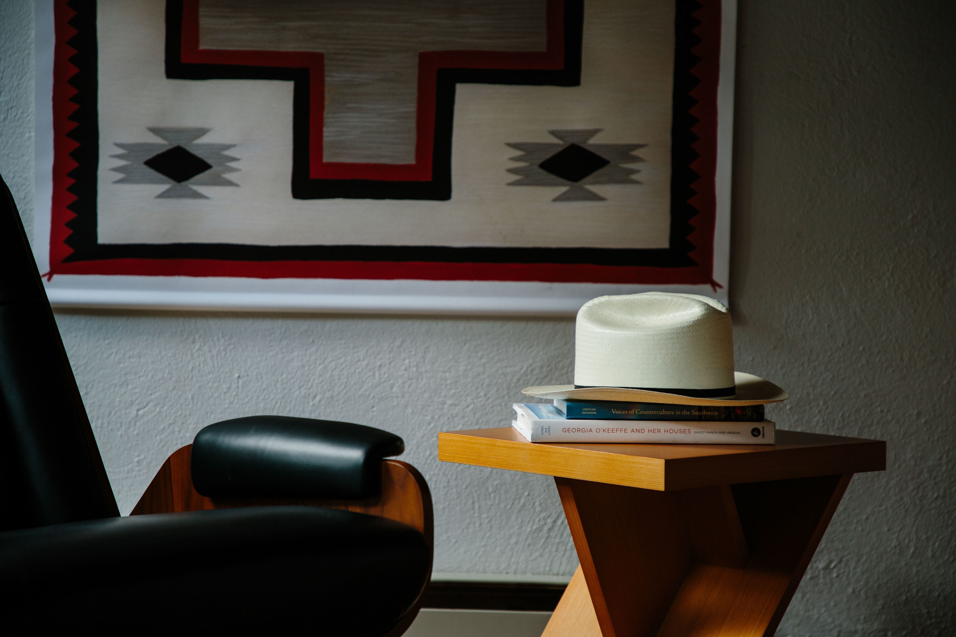 Una habitación con una mesita de madera que sostiene un sombrero y libros, una silla negra y una pared con un tapiz de diseño geométrico.