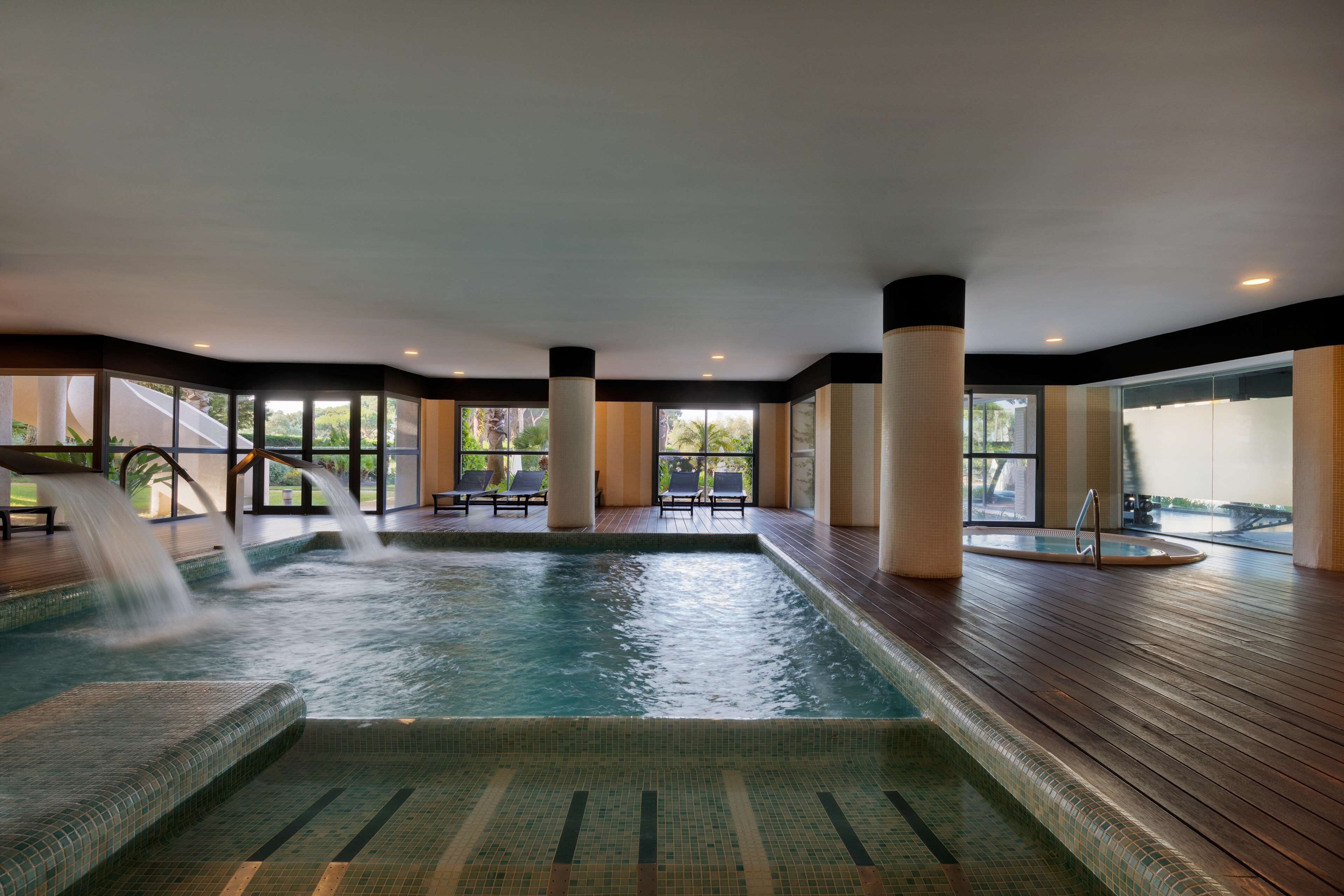 Une piscine intérieure dotée d’une terrasse en bois, de chaises longues et d’un élément décoratif aquatique.