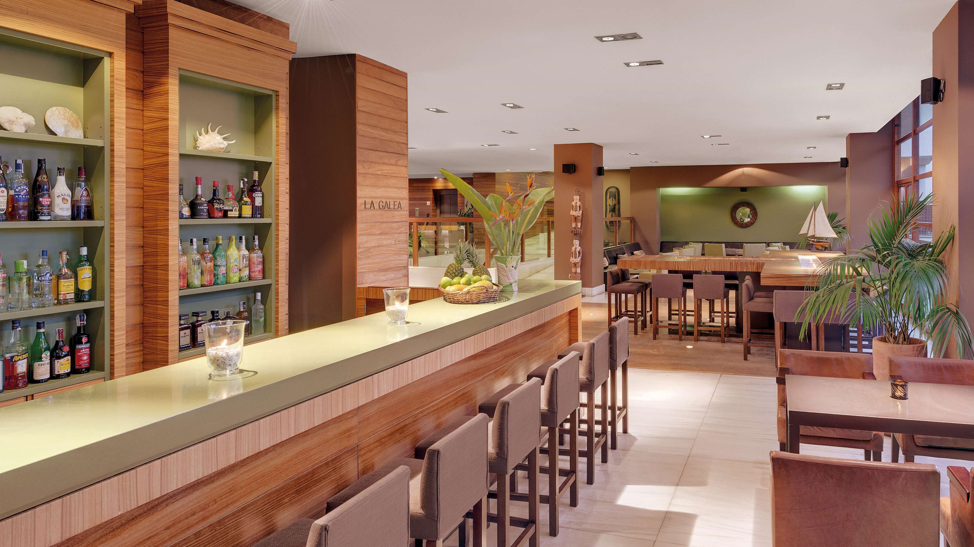 A bar area with wooden shelves stocked with various bottles, a fruit basket, and a seating area with chairs and tables.
