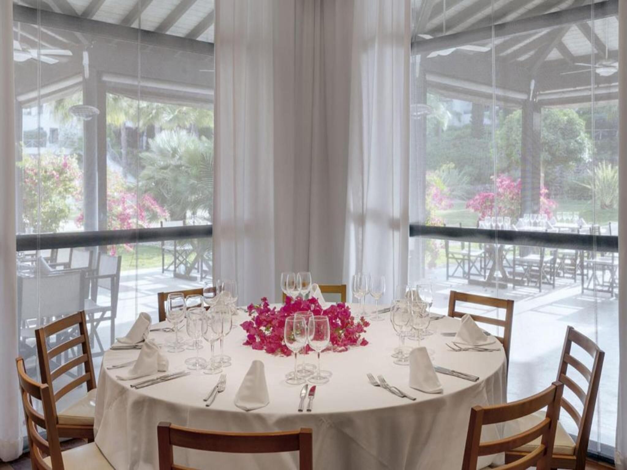 Una mesa redonda dispuesta para comer, con sillas, un mantel blanco y un centro de mesa floral.