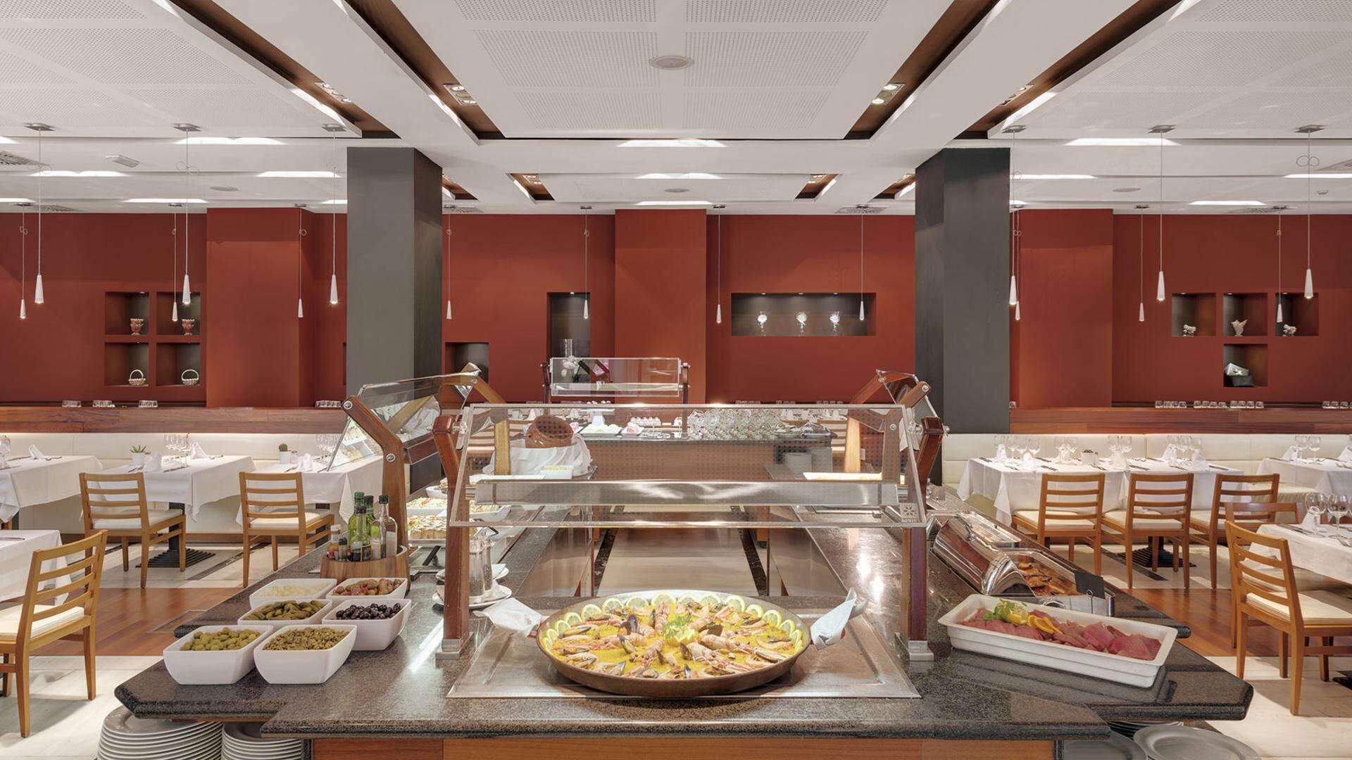 Moderni ruokailutila, jossa on useita pöytiä, keskeisellä paikalla oleva buffetpöytä ja monipuolinen valikoima erilaisia ruokia esillä.