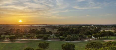 Campo de golf con un camino despejado, árboles y un atardecer de fondo.