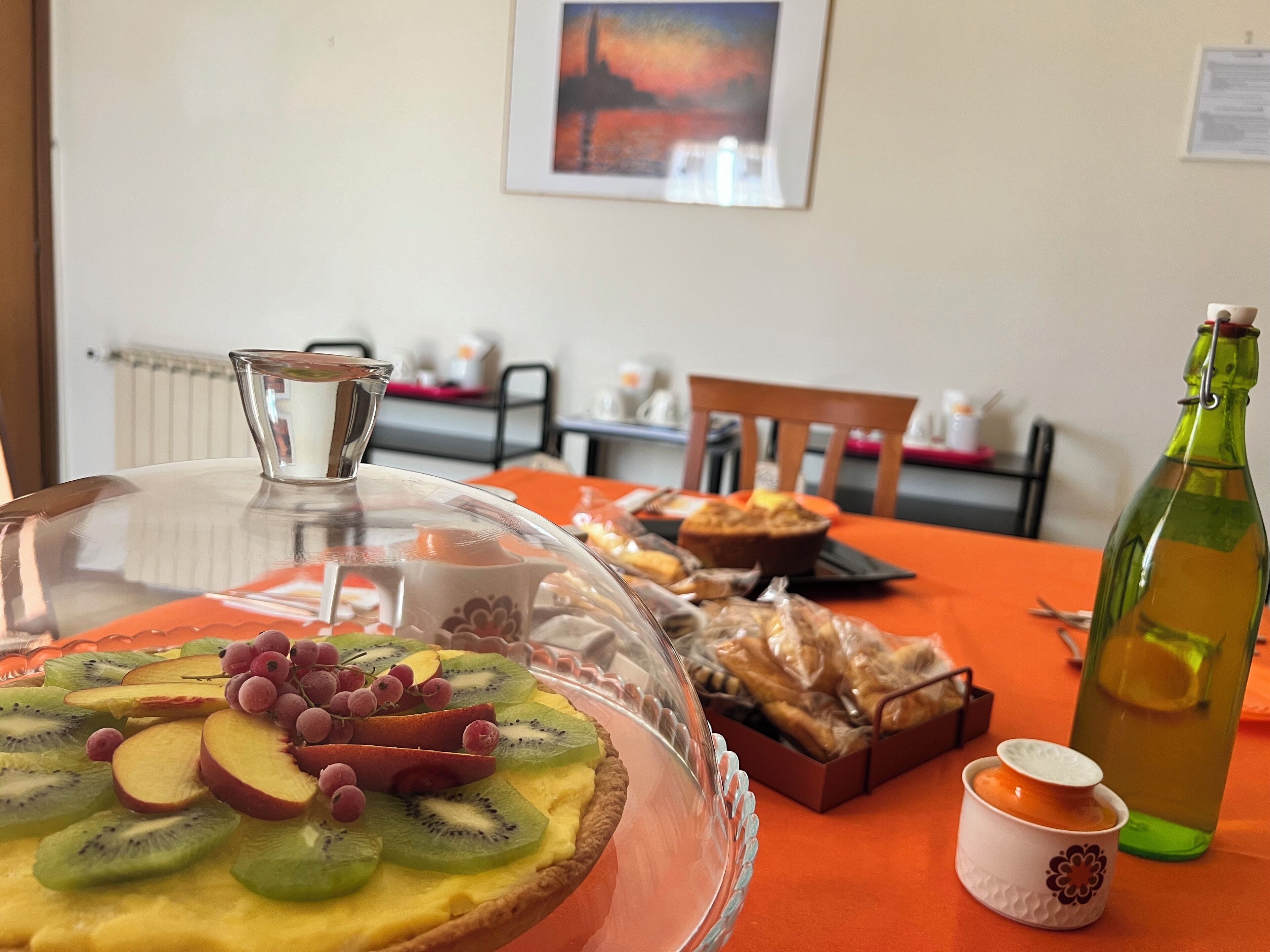 A table with a covered fruit tart, a bottle of olive oil, and a small container, likely for condiments, on an orange tablecloth.