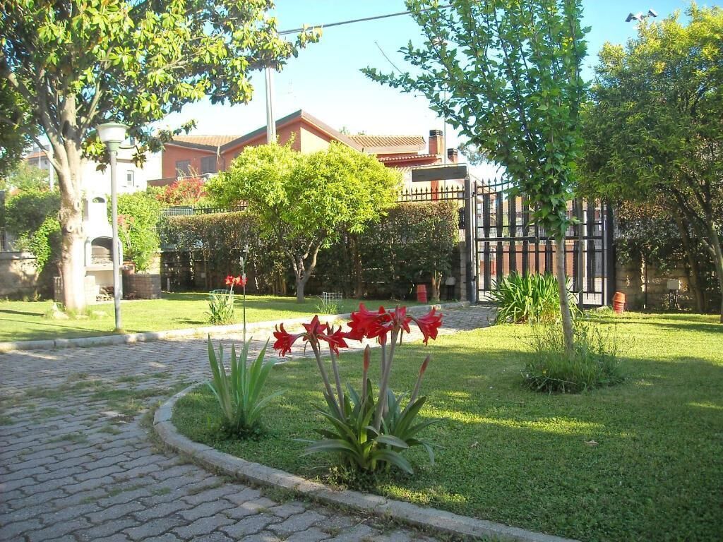 A courtyard with a paved walkway, a small garden with red flowers, and a building with a red roof in the background.