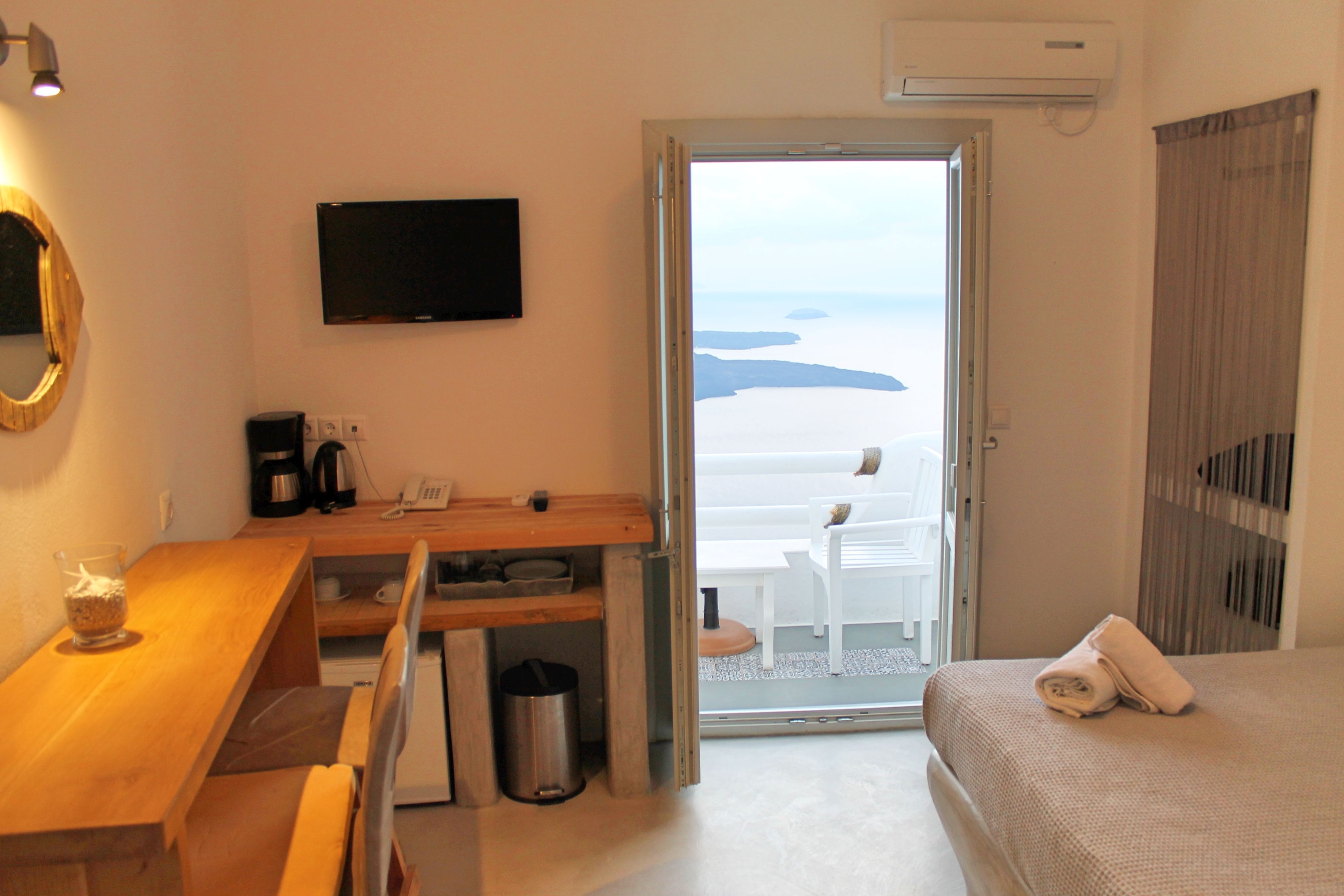 A hotel room with a wooden desk, a television, a bed with a rolled towel, and a view of the beach through an open door.